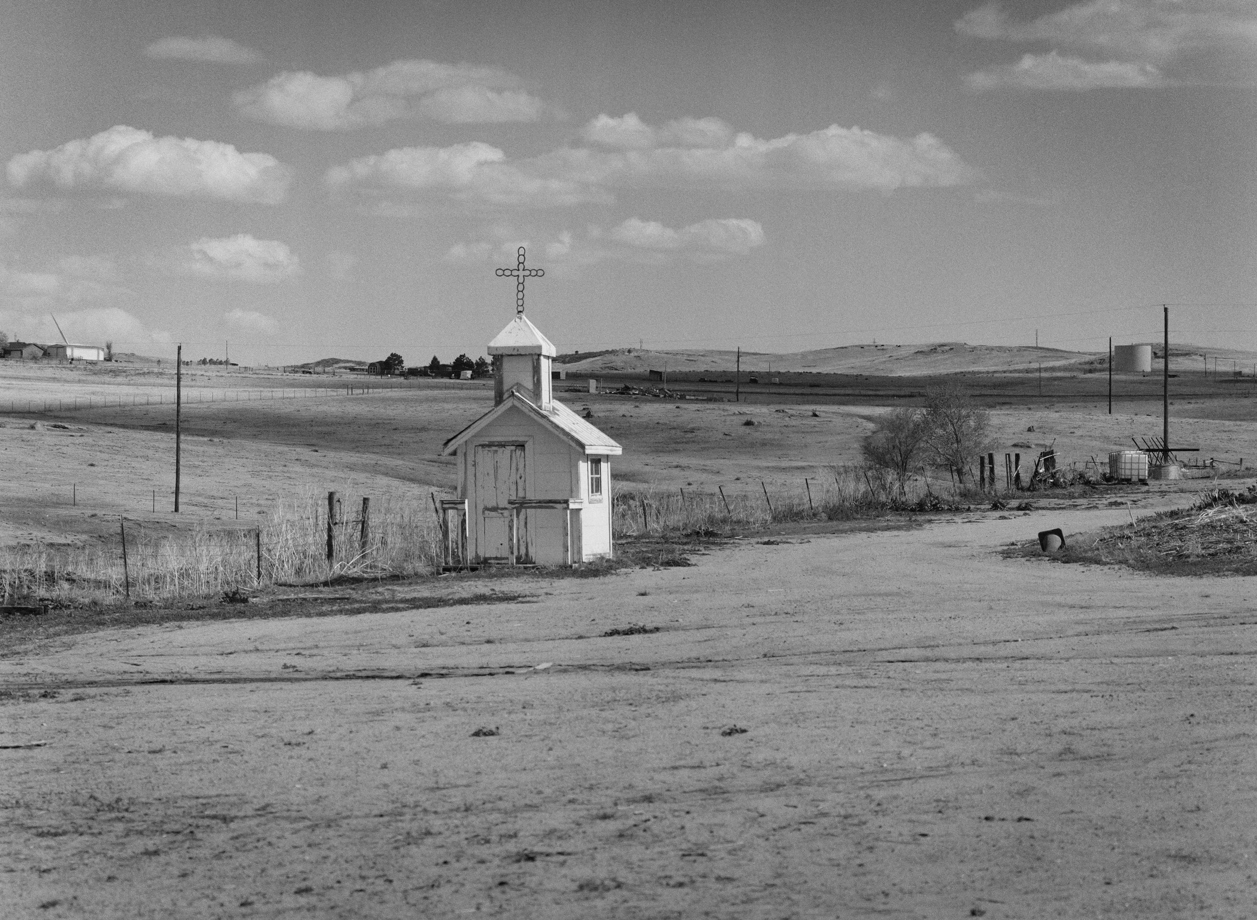 A small rural church with a cross on its steeple, standing in an open, dusty landscape with scattered trees and power lines, under a partly cloudy sky.