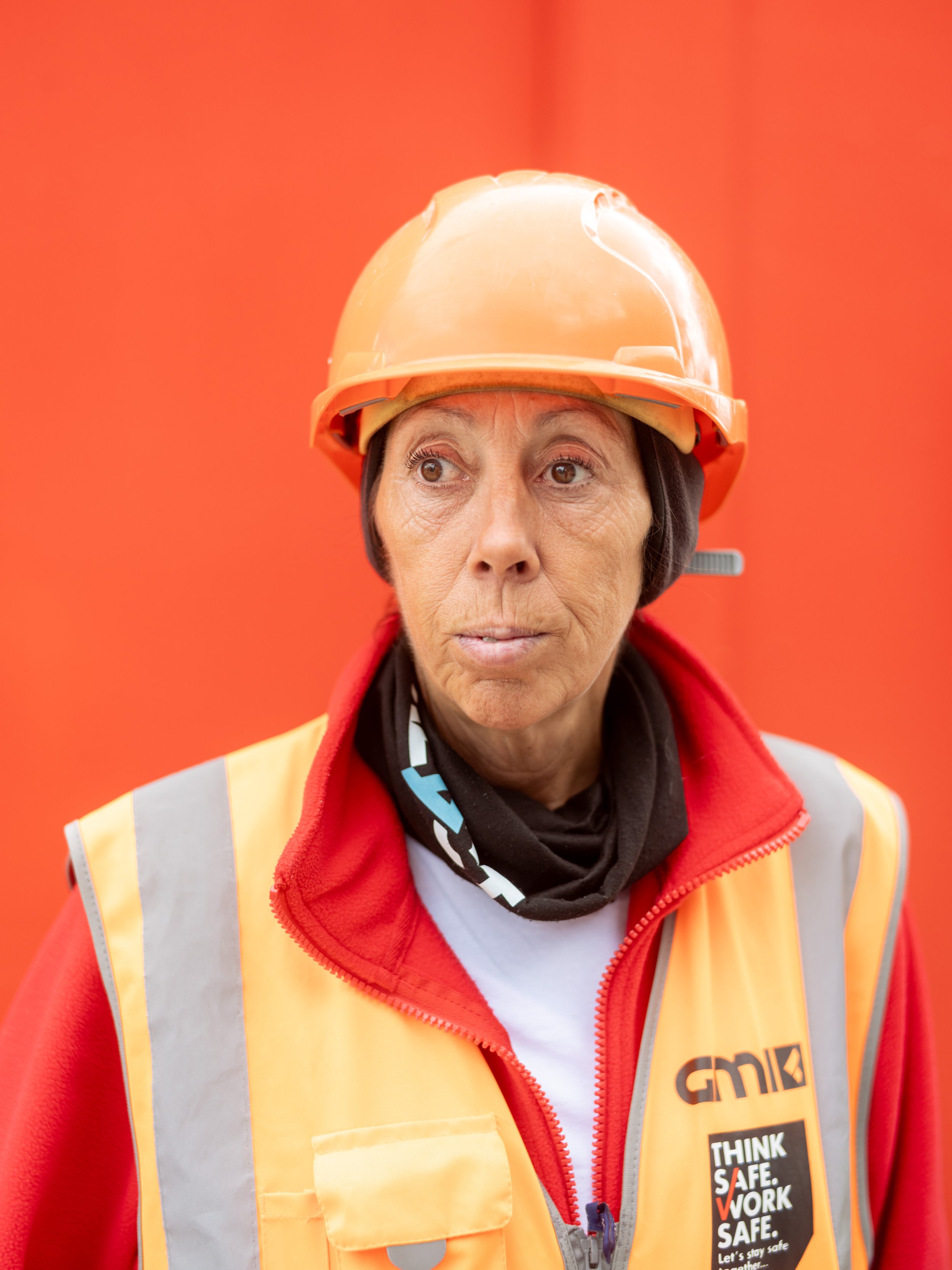 A woman wearing a construction orange helmet and safety vest, with a black neck gaiter, standing against a solid orange background.