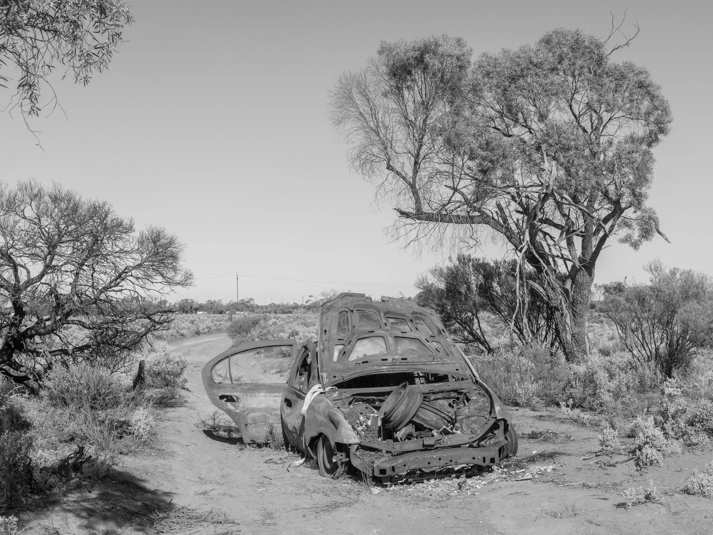 A black and white photograph of a wrecked, abandoned car in a desert landscape with sparse bushes and trees, and a trail in the background.