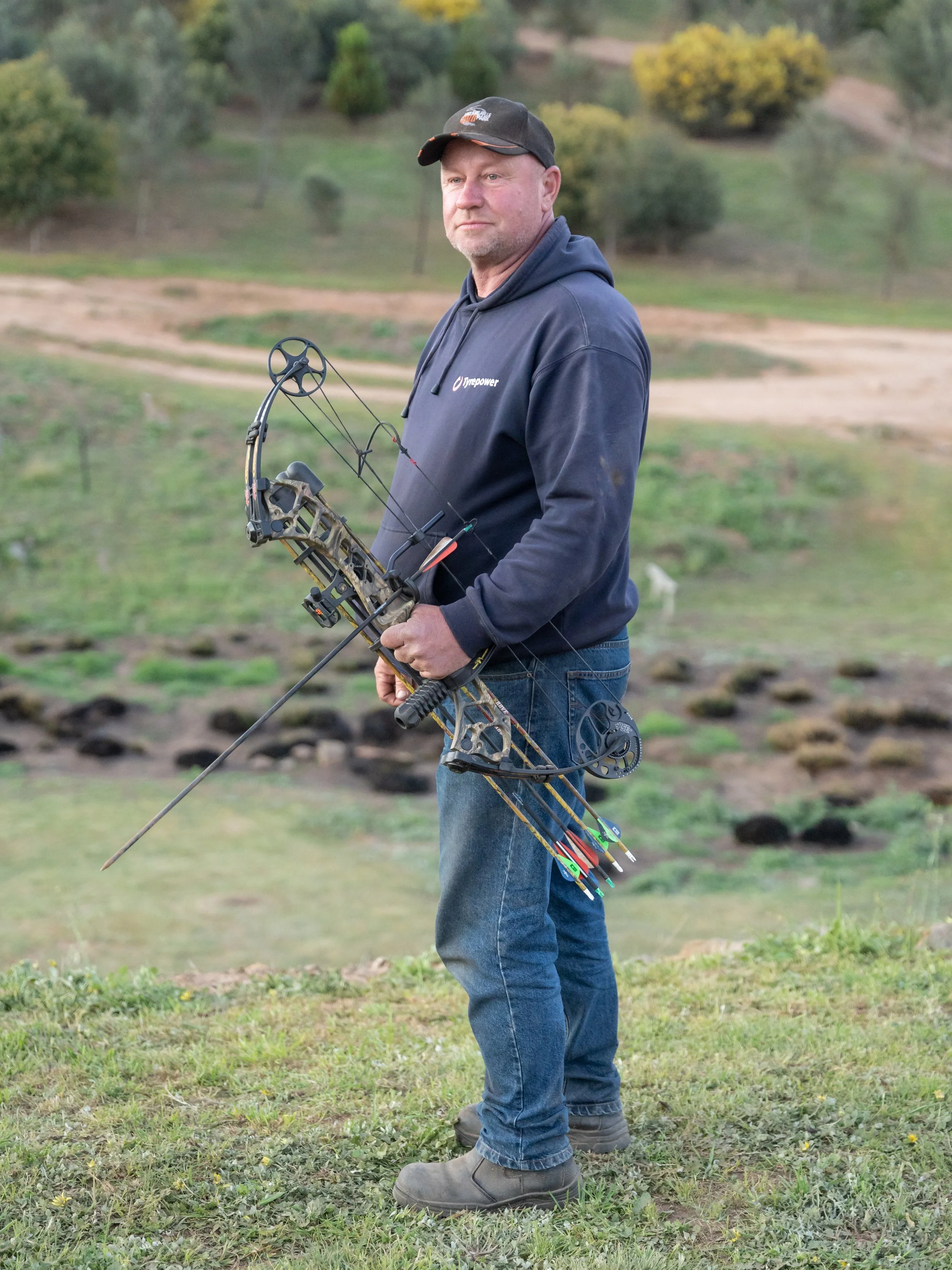 A man in a hoodie and jeans holding a compound bow with arrows, standing outdoors in a grassy area with trees and dirt pathways in the background.
