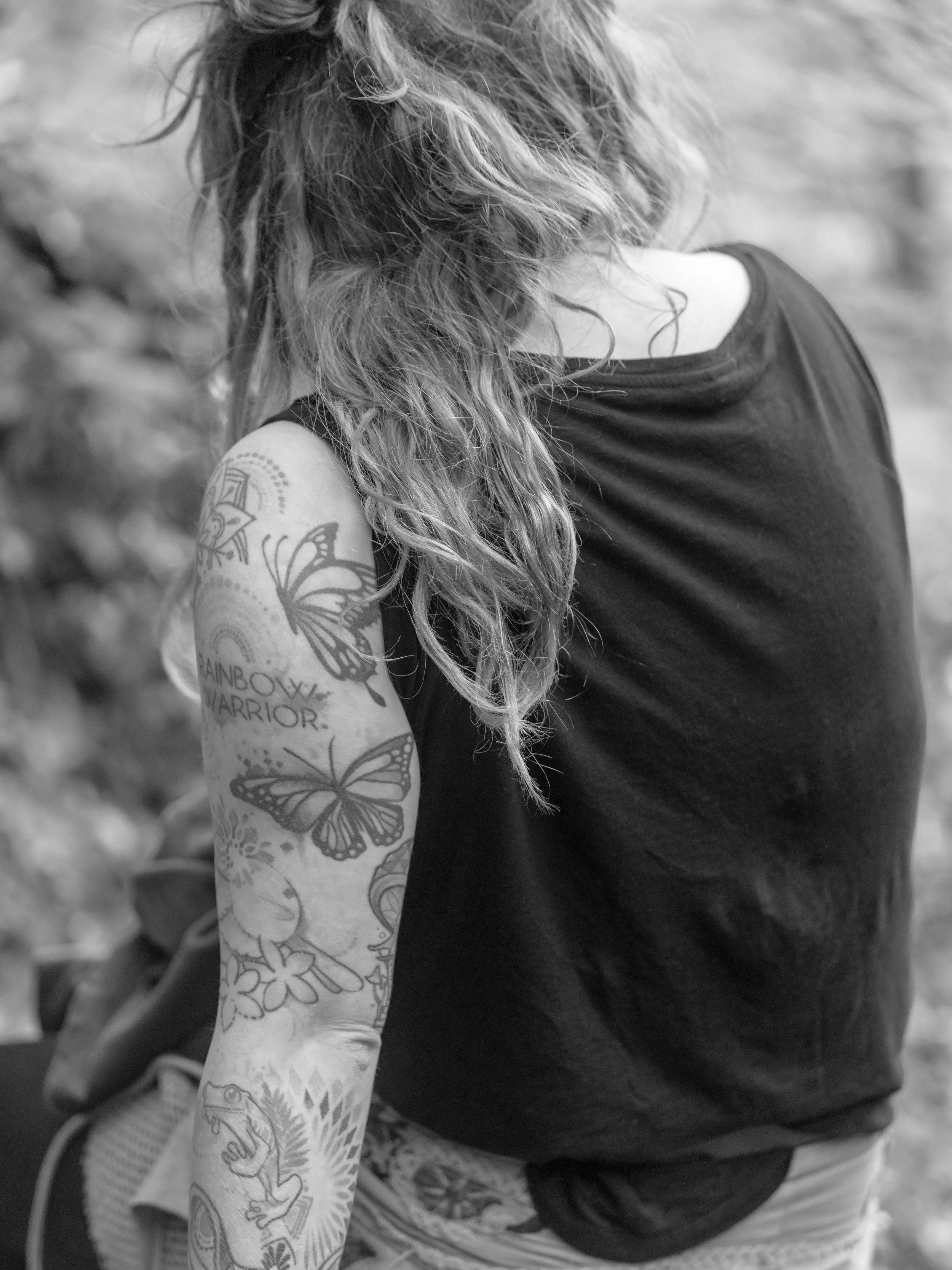A woman with curly hair wearing a sleeveless black top, showing her tattooed arm with butterflies, a chameleon, and other designs.