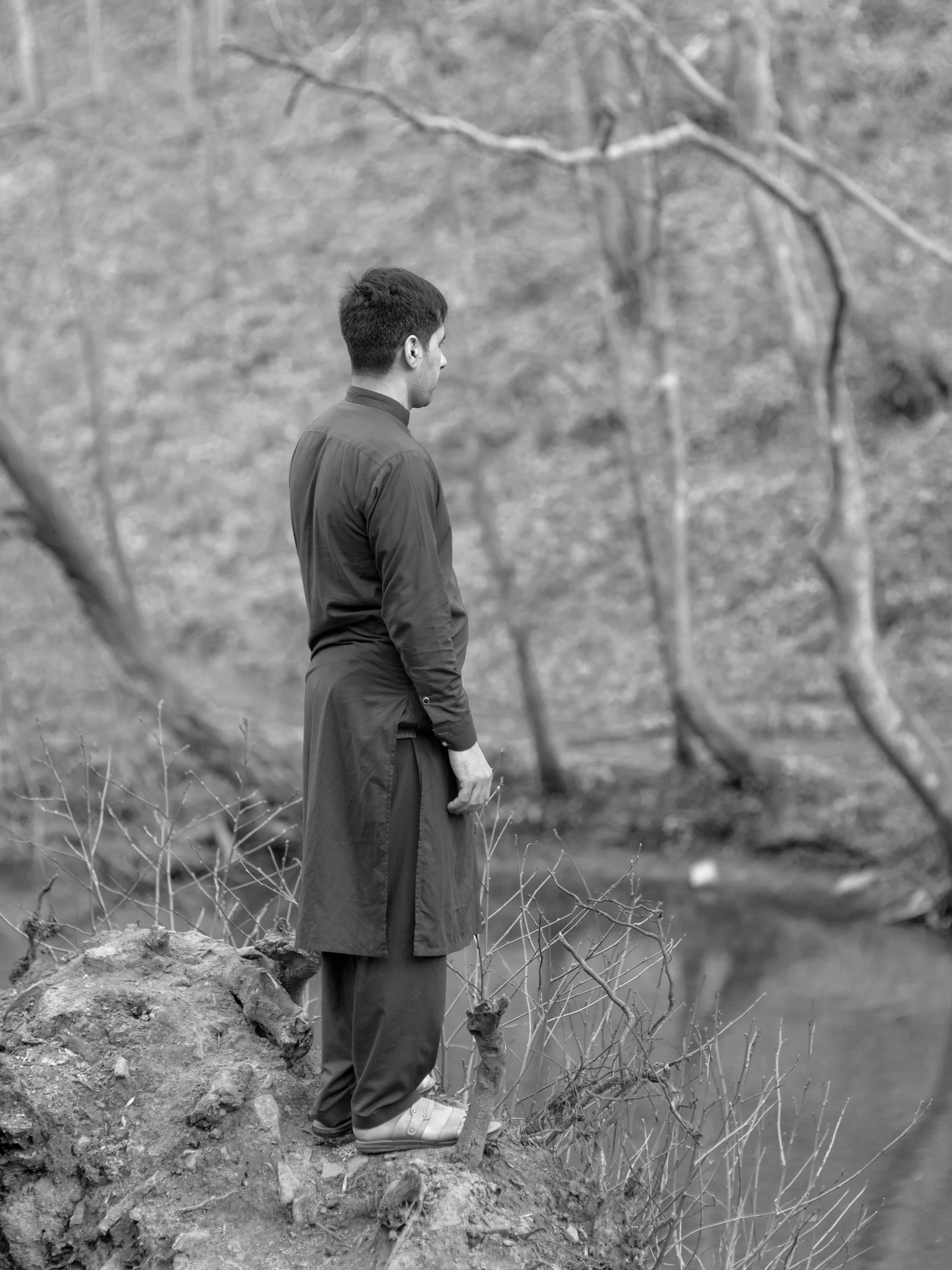 A man stands on a rocky outcrop near a small creek in a bare, wooded area, looking into the water.