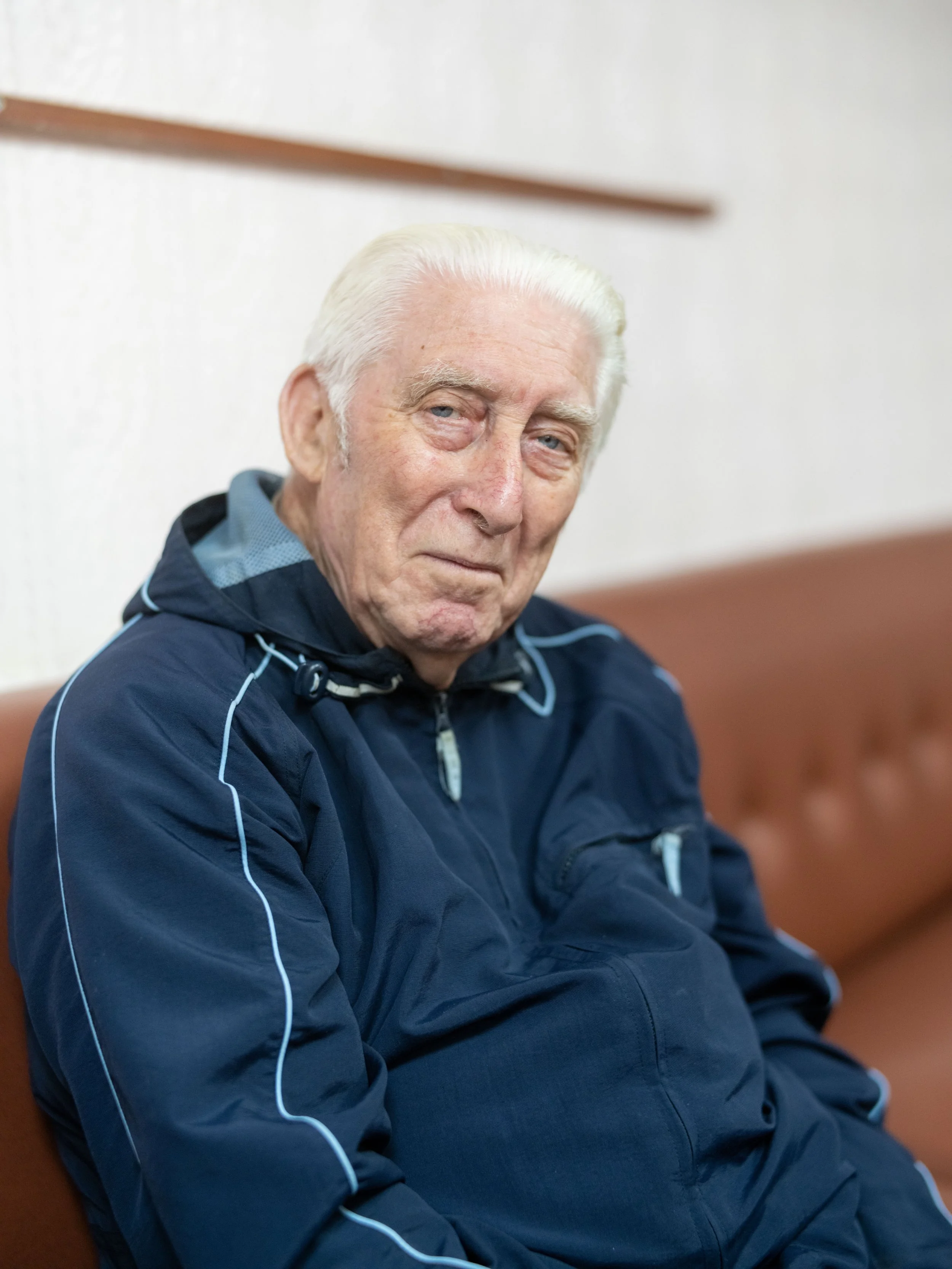 An elderly man with white hair and a solemn expression sitting on a brown couch, wearing a blue jacket with light blue piping.