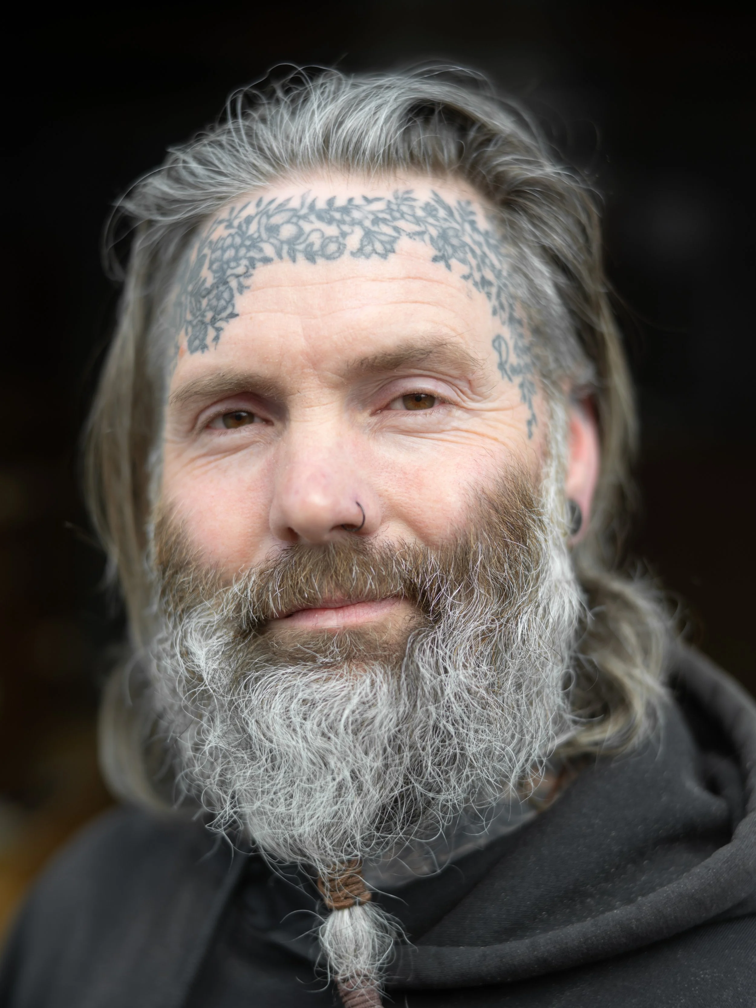 Close-up of a smiling man with gray hair, a thick beard, a nose piercing, and a tattoo on his forehead.