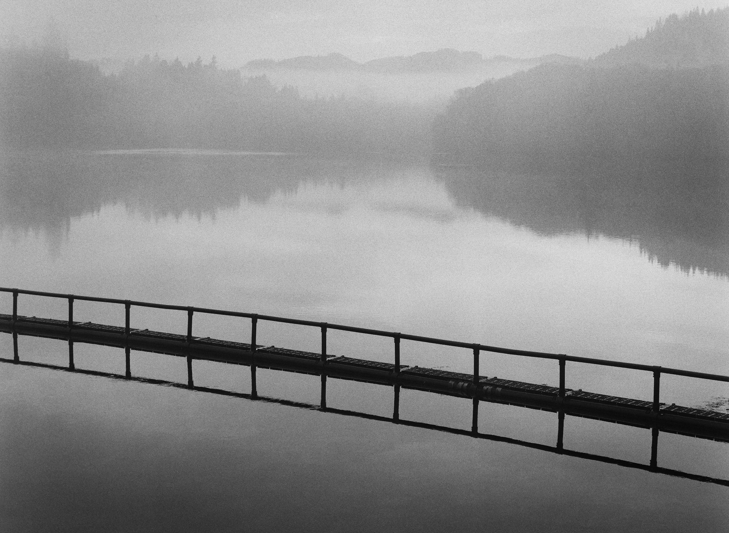 A black and white photo of a calm lake with a walkway in the foreground and a mirror-like reflection of the surrounding mountains and trees in the water.