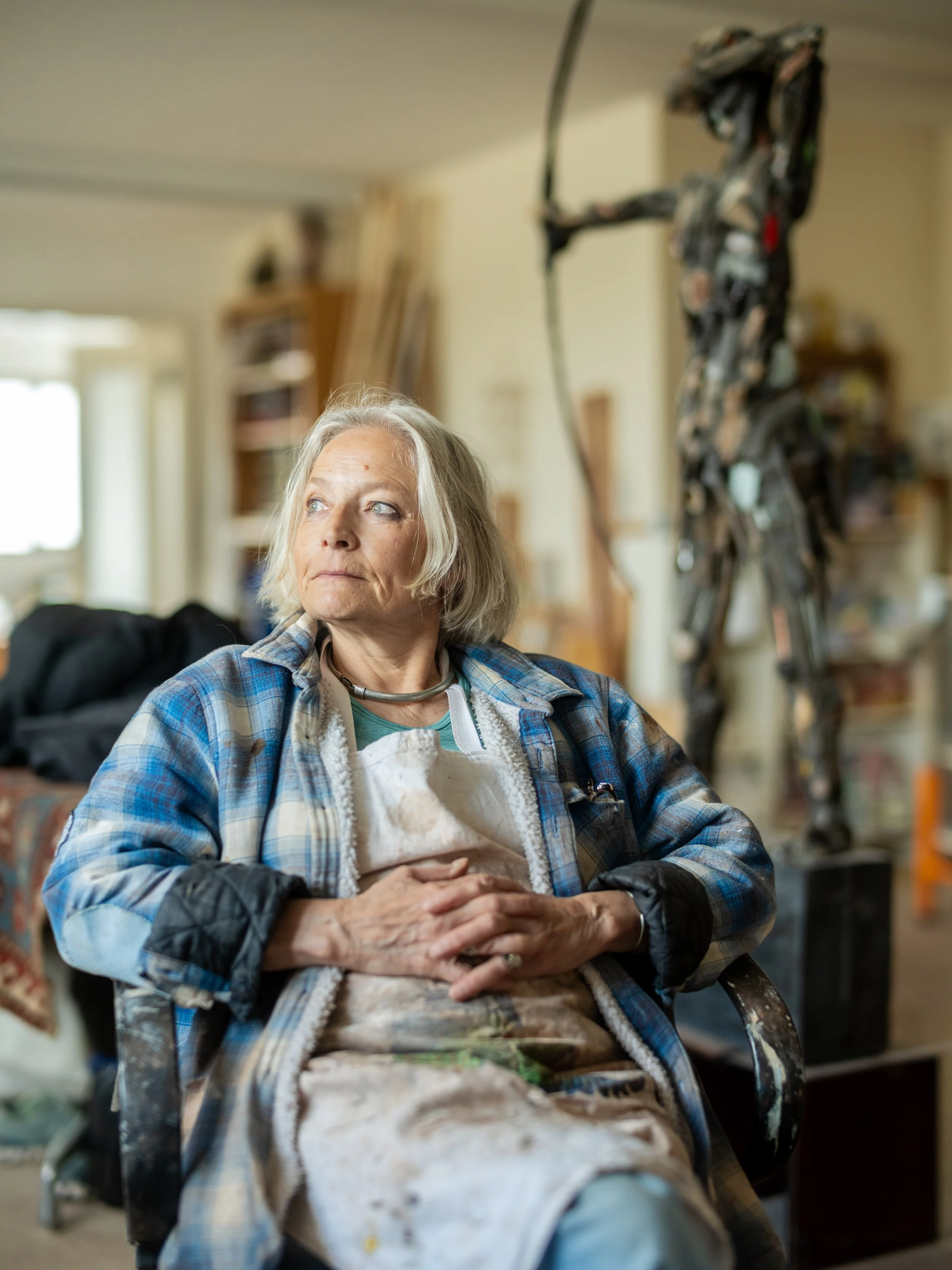 Older woman with gray hair sitting on a chair in an art studio, surrounded by artwork and supplies, with a large abstract sculpture in the background.