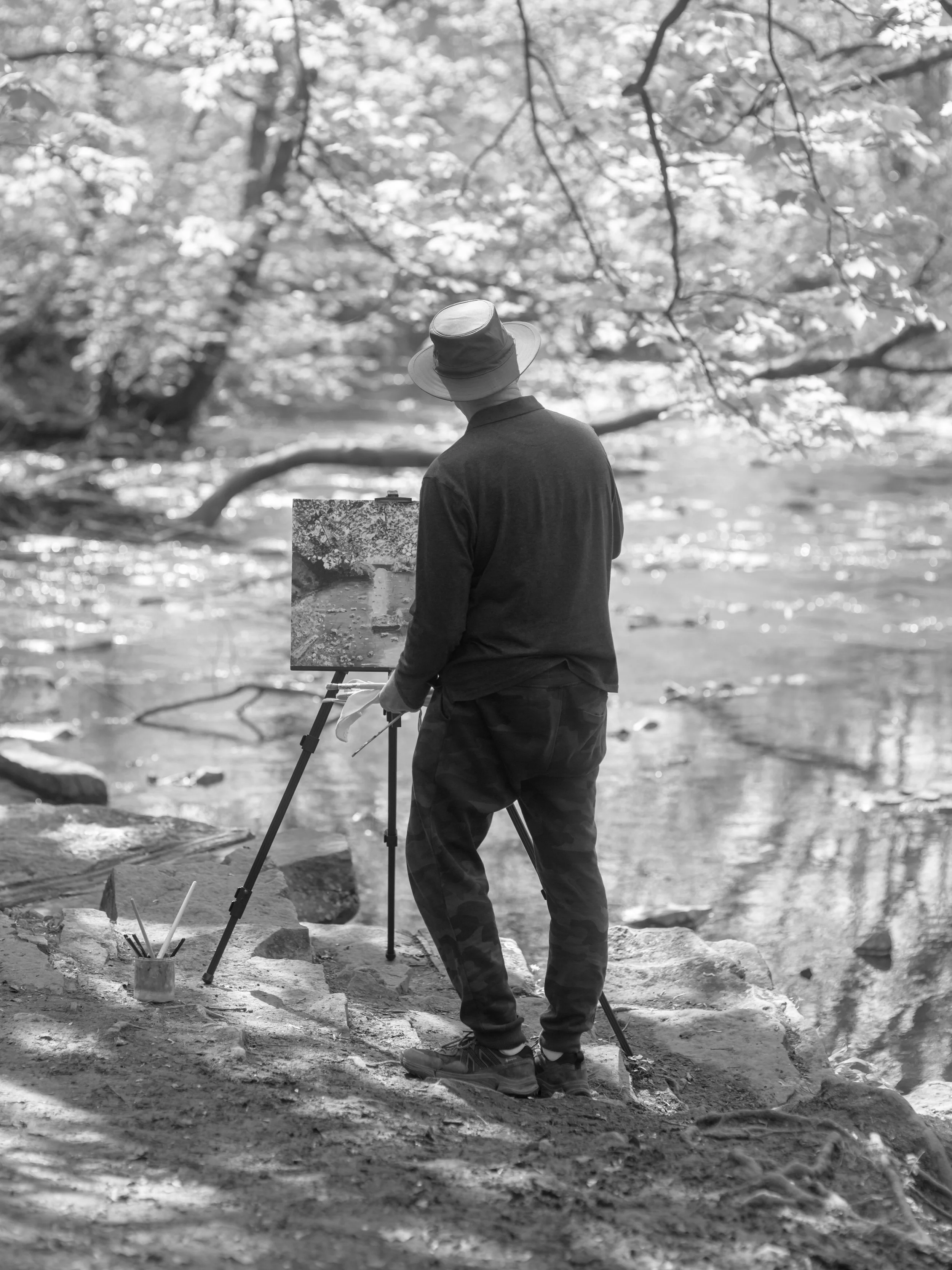 A person standing by a river, painting a landscape on an easel. The scene is outdoors surrounded by trees and rocks, with the person wearing a hat and casual clothing.