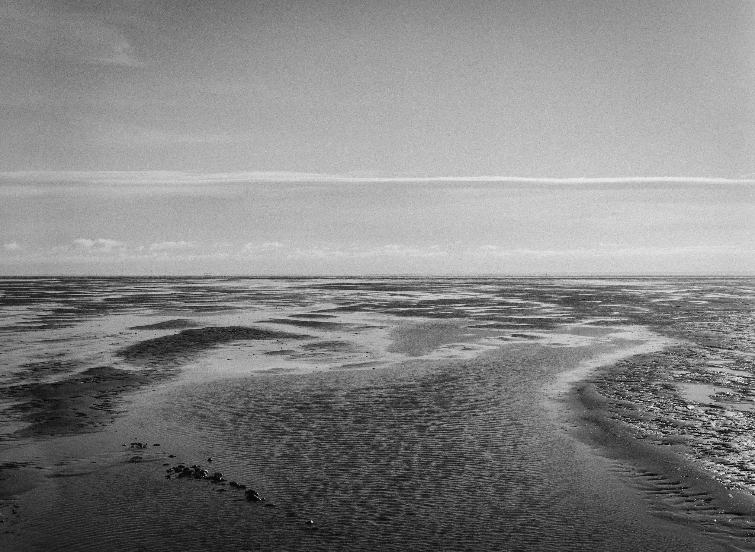 Black and white photo of a sandy beach with shallow water and small pools, extending to the horizon under a sky with clouds.
