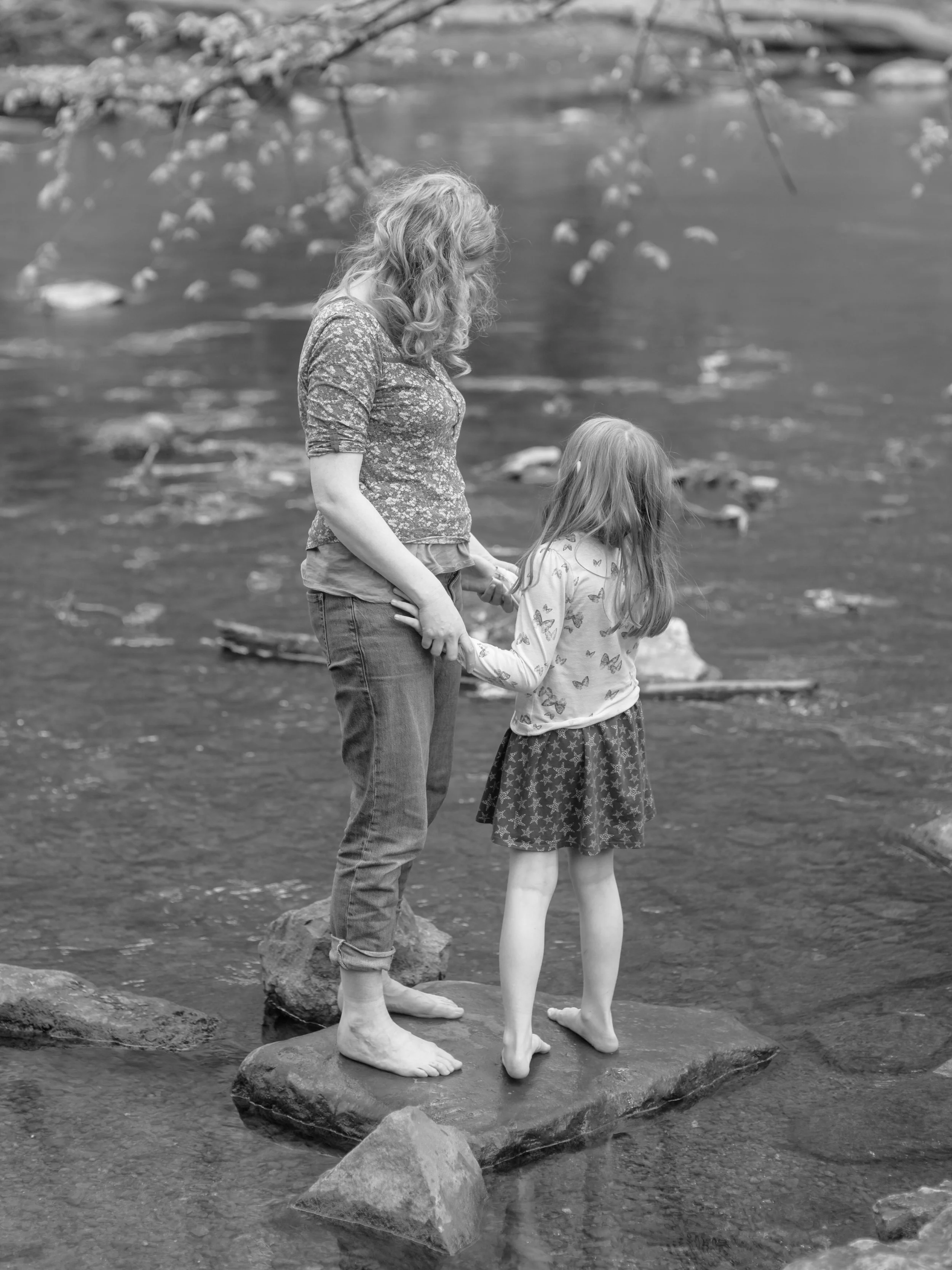 A woman and a young girl holding hands and standing barefoot on a large rock in a river.