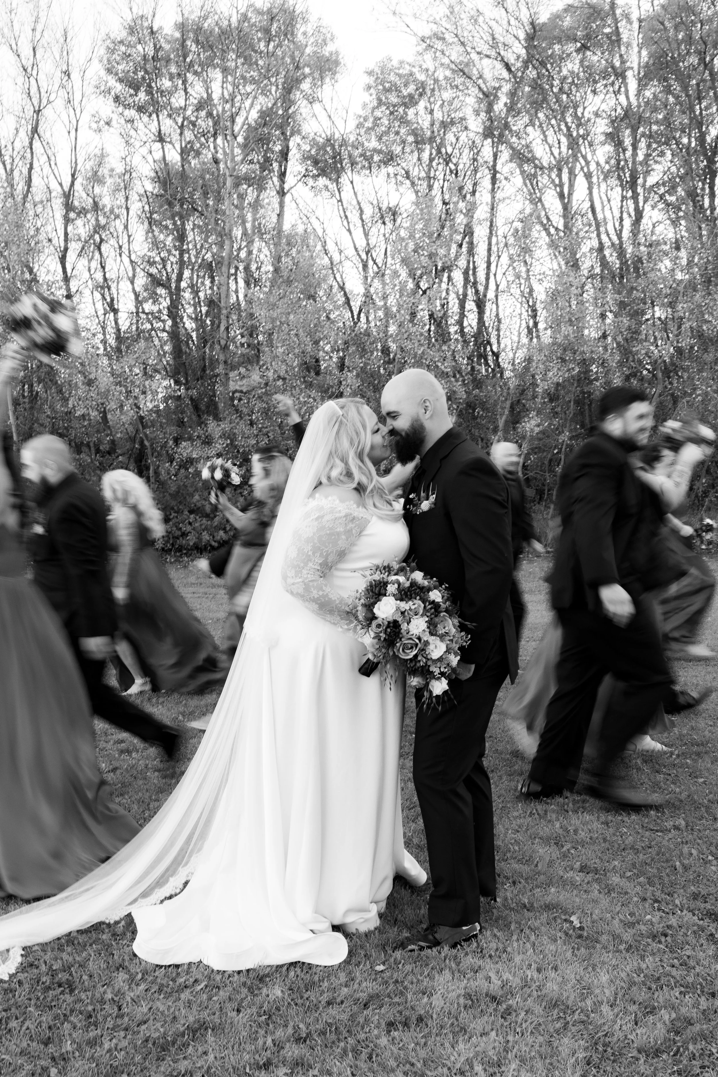 A black and white photo of a couple in wedding attire, smiling and leaning close together, surrounded by blurred wedding guests walking by in an outdoor setting with trees in the background.