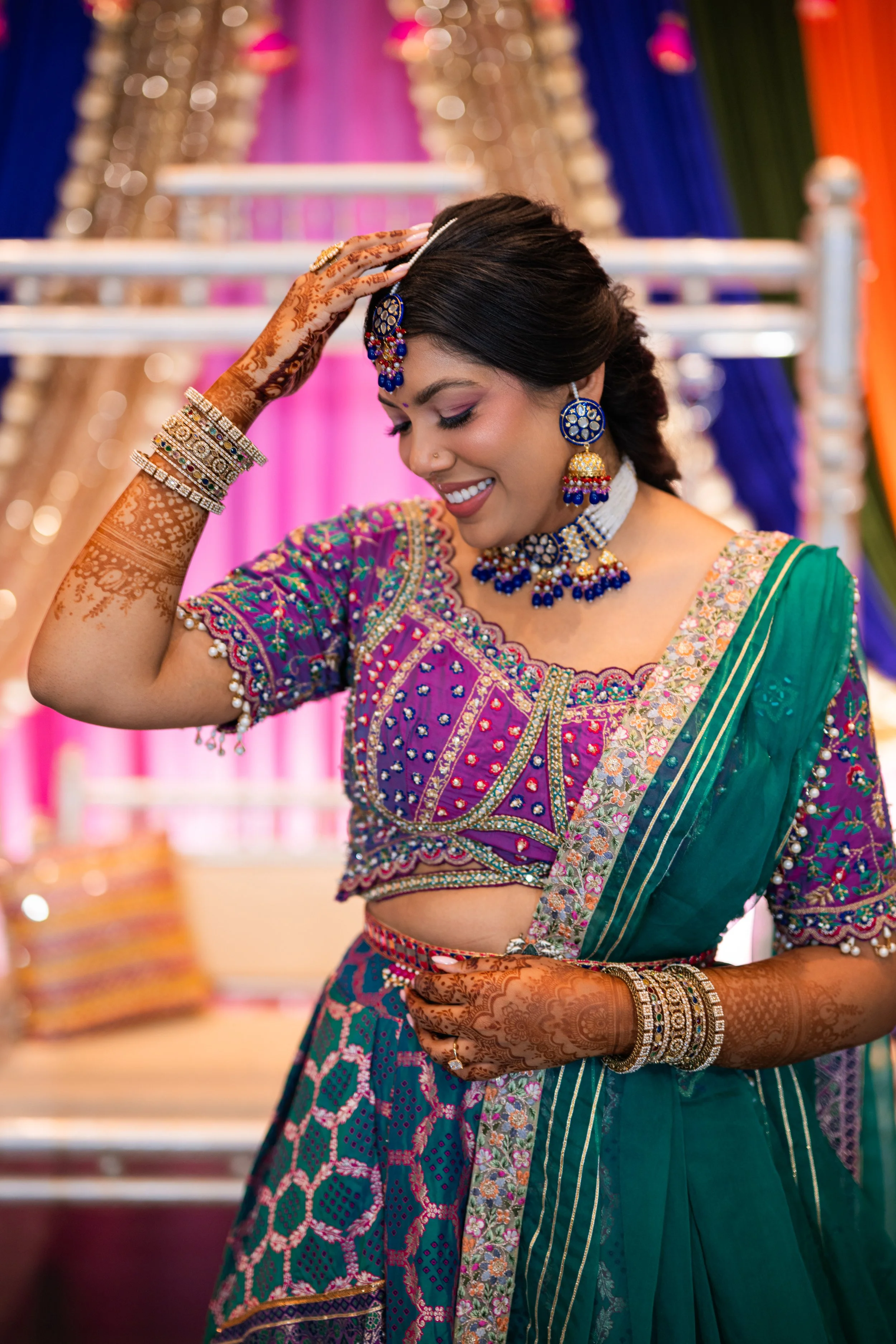 A woman dressed in traditional Indian attire is smiling and adjusting her hair. She is wearing jewelry including earrings, a necklace, bangles, and a headpiece. She has henna designs on her hands and arms. The background features colorful drapes in p
