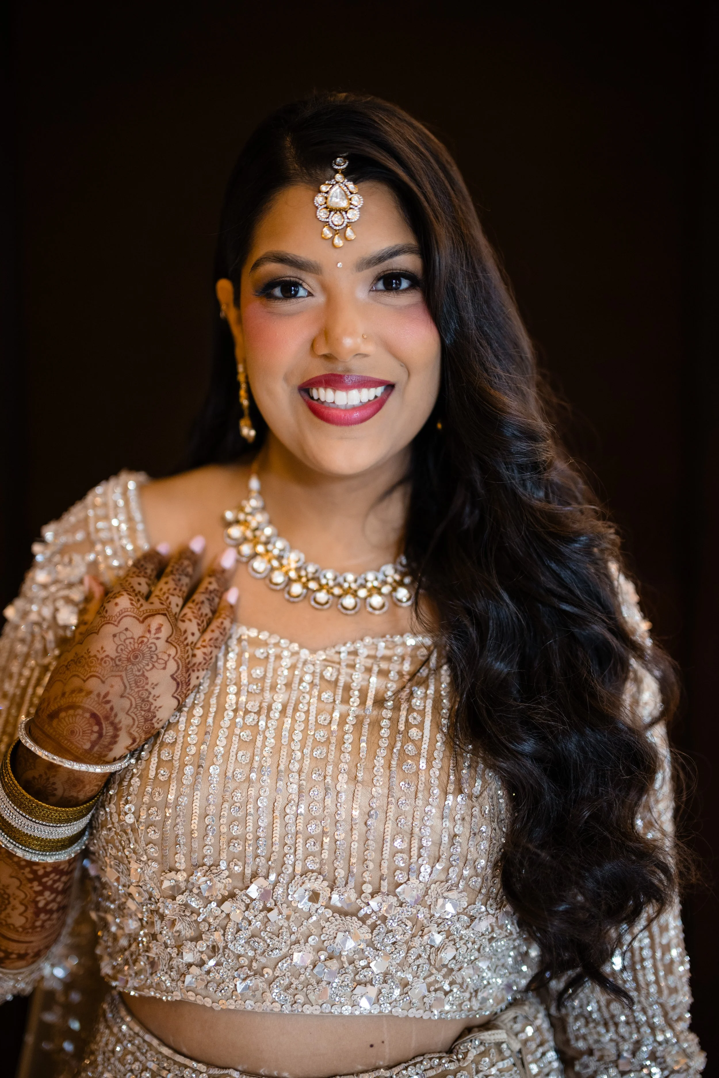 A woman dressed in traditional Indian bridal attire, wearing ornate jewelry including earrings, necklace, and a maang tikka, with intricate henna on her hand, smiling against a dark background.
