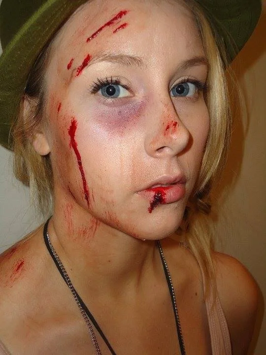 Young woman with blonde hair wearing a green hat and a necklace, with fake blood and makeup wounds on her face and shoulder, and a bruised eye.