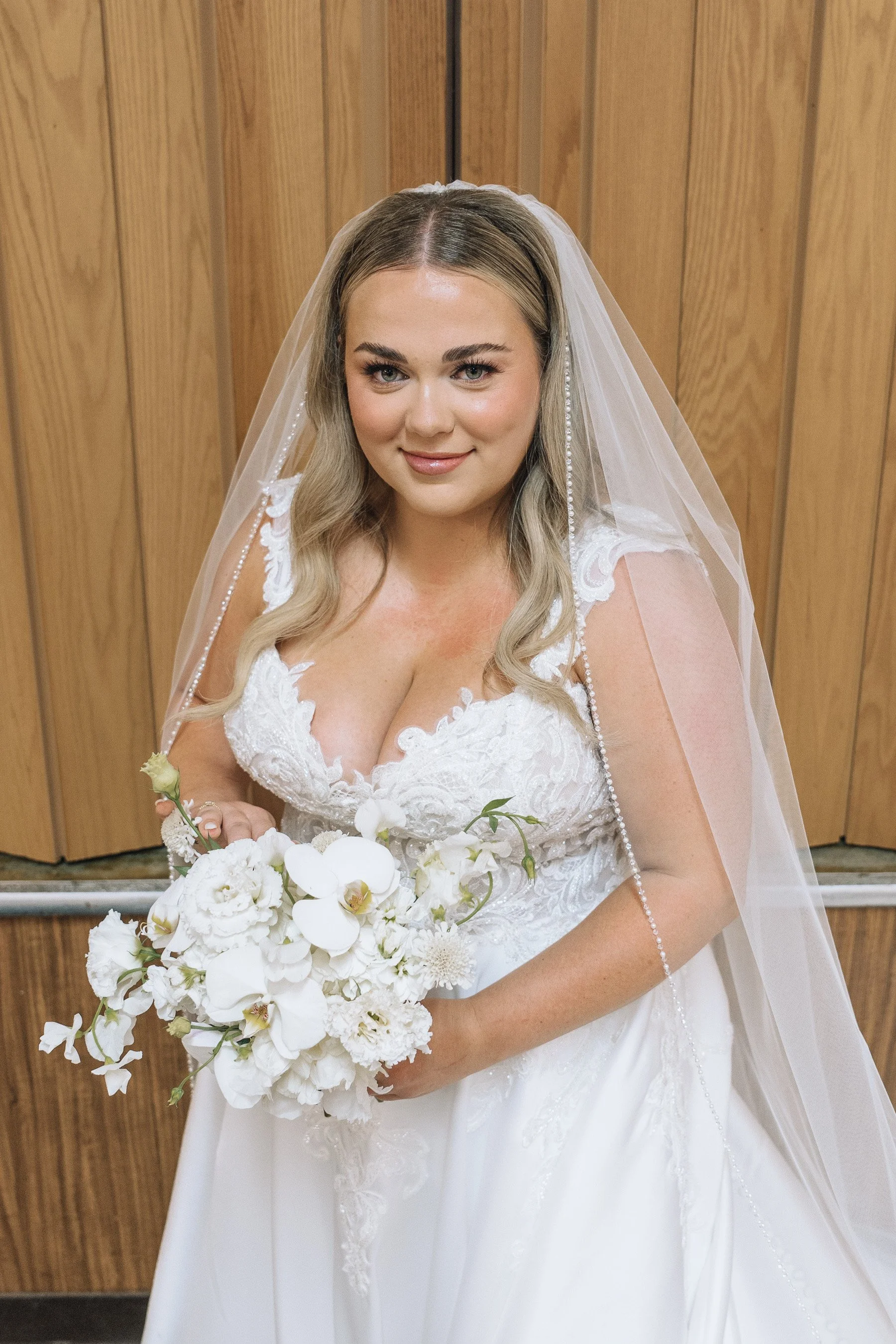 A bride in a white wedding dress holding a bouquet of white flowers, standing in front of a wooden wall.