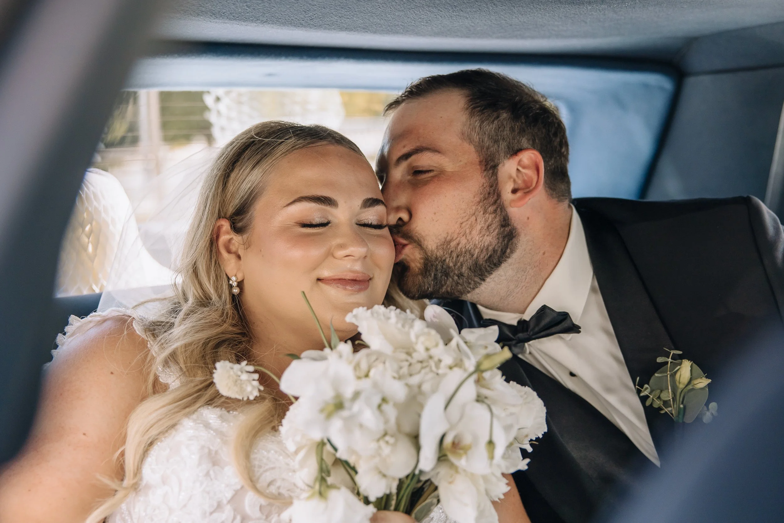 Bride and groom sharing a kiss in the back of a car during their wedding, with the bride holding a bouquet of white flowers.