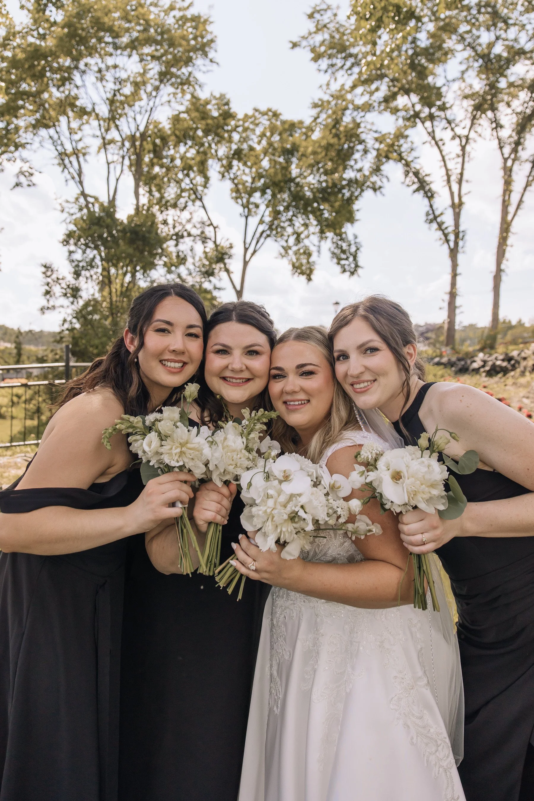 A group of four smiling women in black dresses and a bride in a white lace wedding gown holding bouquets of white flowers, in an outdoor setting with trees and a cloudy sky in the background.