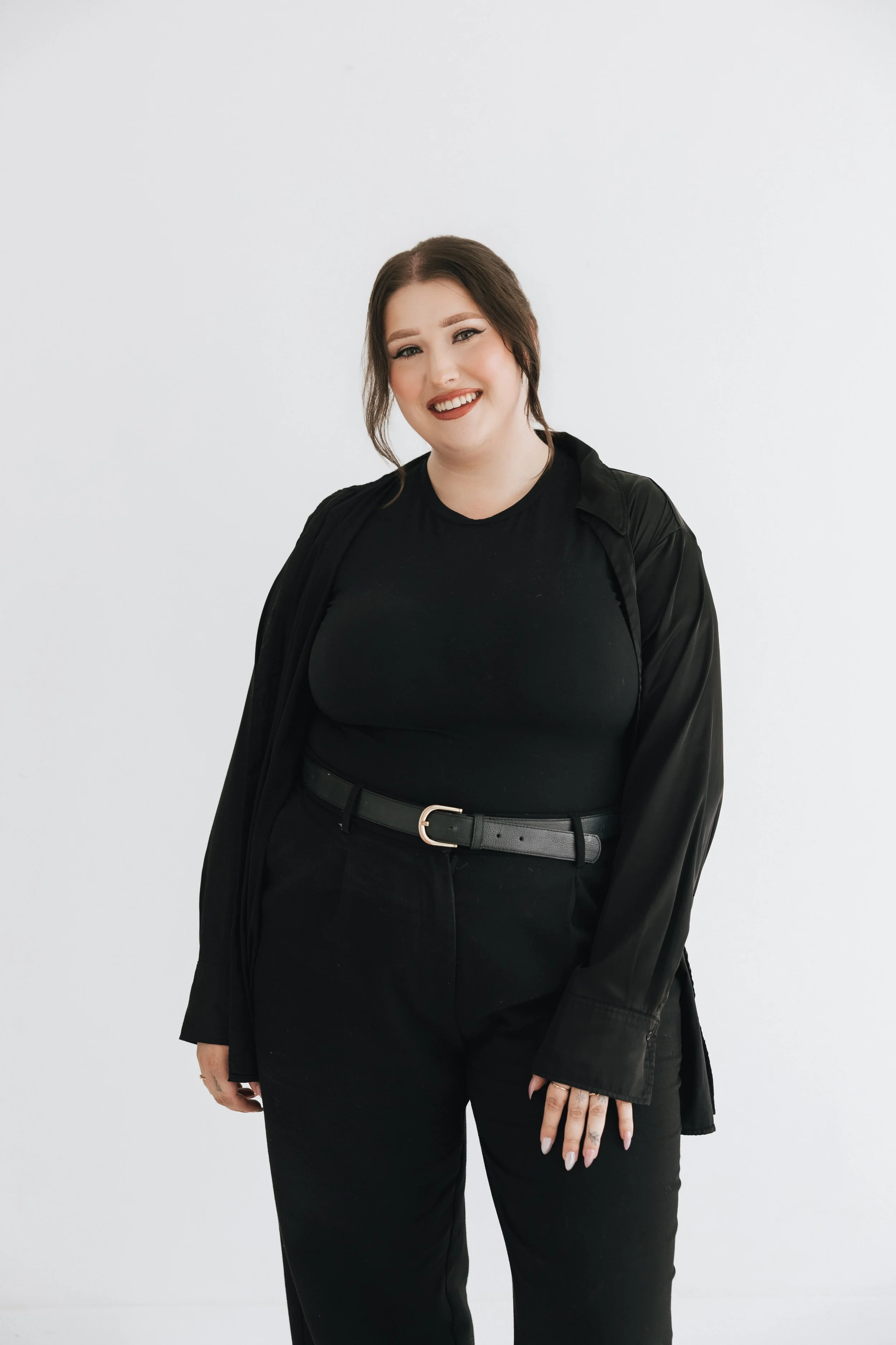 A woman with shoulder-length brown hair styled in loose waves, wearing a black top, black pants, and a black satin jacket draped over her shoulders, standing against a plain white wall, smiling at the camera.