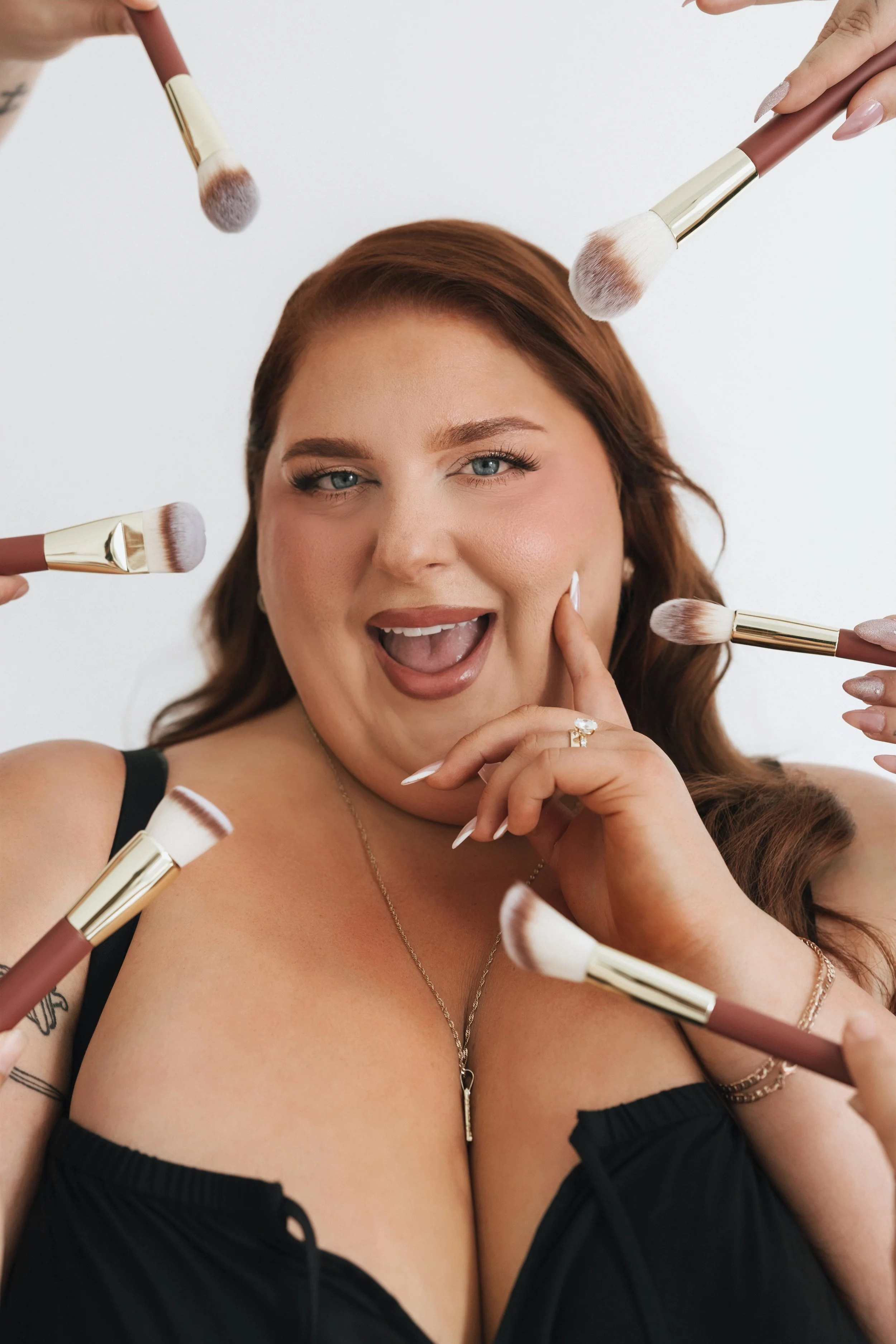 A woman with red hair and blue eyes surrounded by makeup brushes, touching her chin, wearing a black top with shoulder cutouts, jewelry, and makeup.