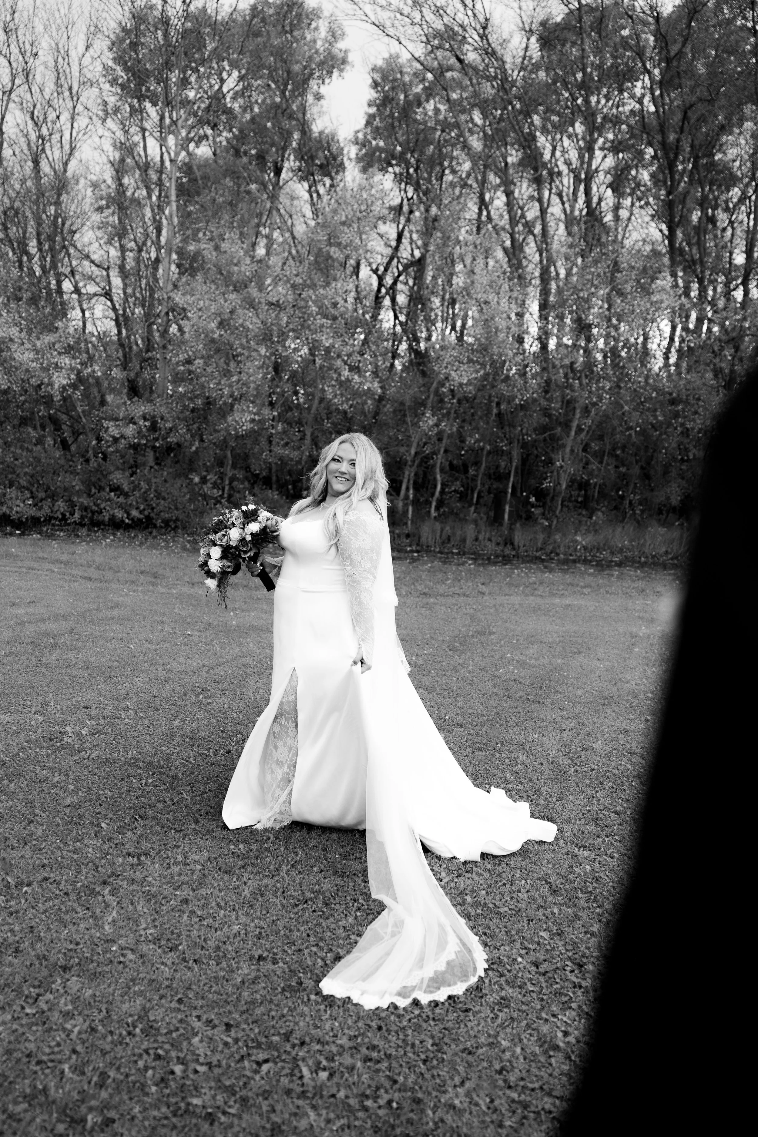 A bride in a long white wedding dress holding a bouquet of flowers, standing outdoors on grass with trees in the background, smiling at the camera.