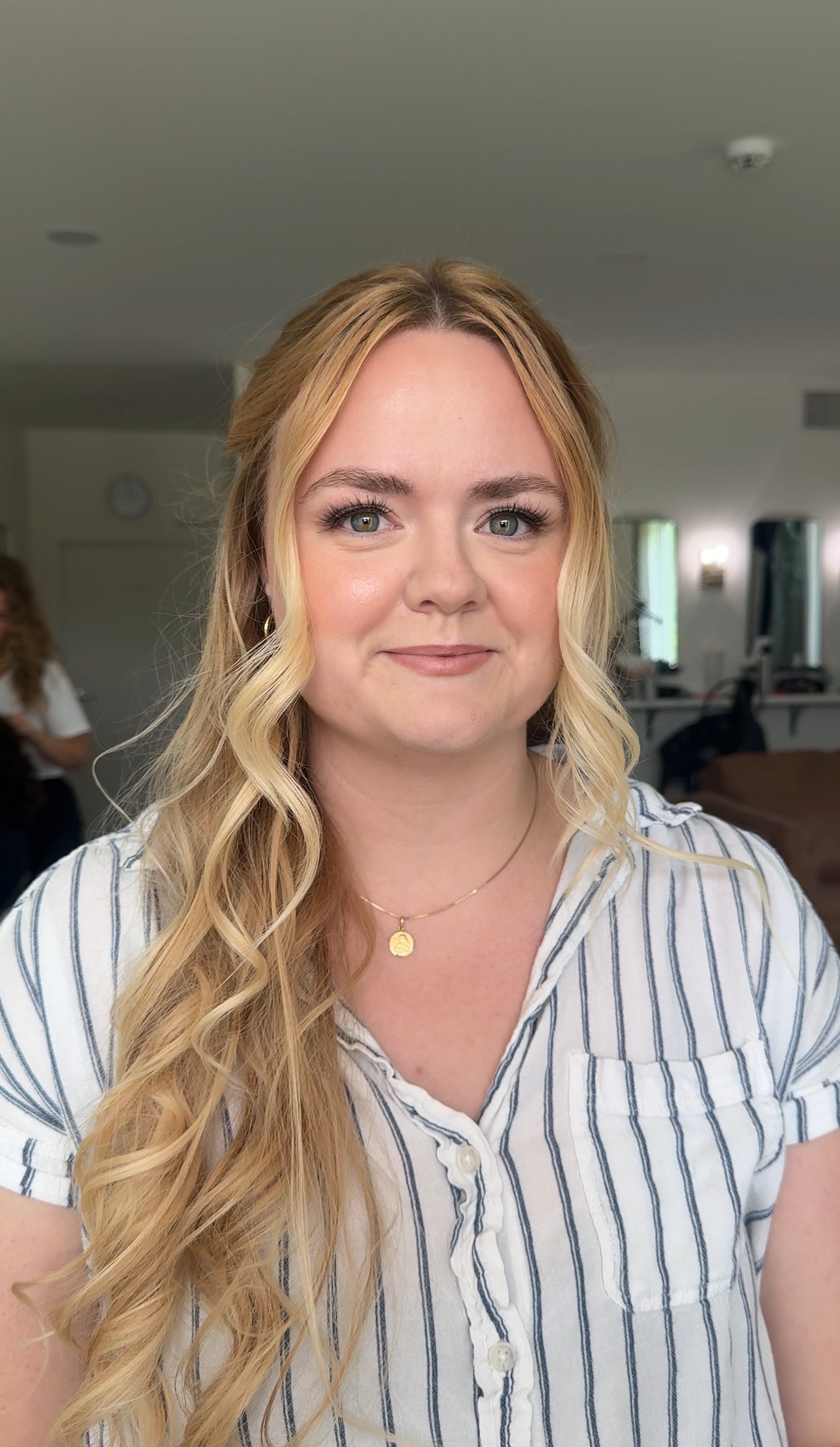 A woman with long, wavy blonde hair, wearing a striped button-up shirt and a gold necklace, smiling in an indoor setting.