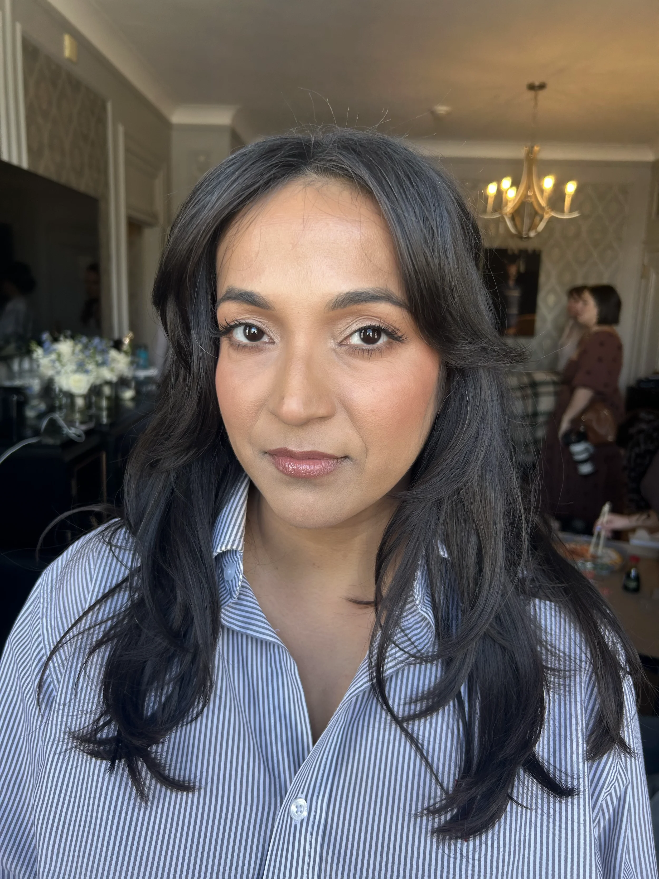 Close-up of a woman with long dark hair and light makeup, wearing a blue and white striped shirt, with a blurred background of an indoor gathering room with people and a chandelier.