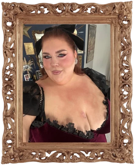 A woman wearing a dark red dress with black lace trim and black puffy sleeves, standing in front of a mirror, with her hair styled and makeup on, inside a decorative gold frame.