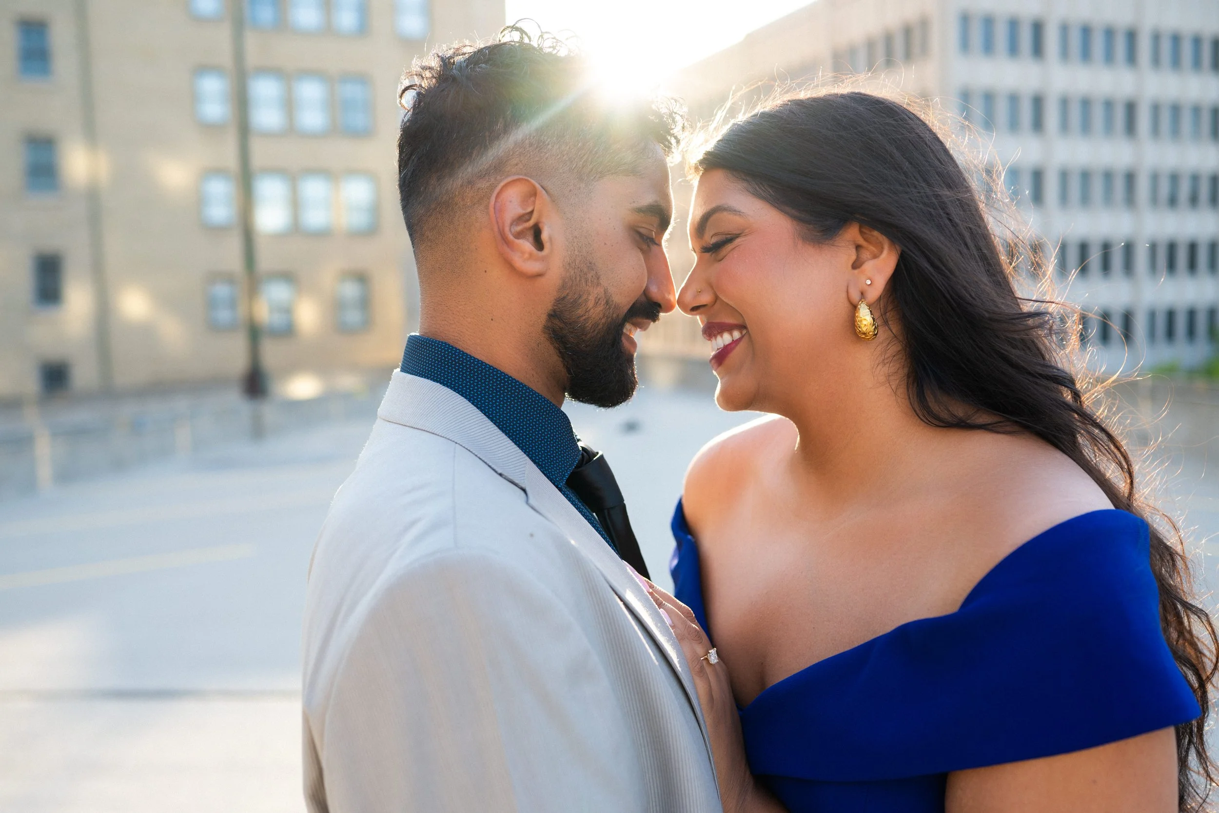 A man and woman are standing close together with their foreheads touching outdoors, smiling with eyes closed, sunlight in the background, buildings around.