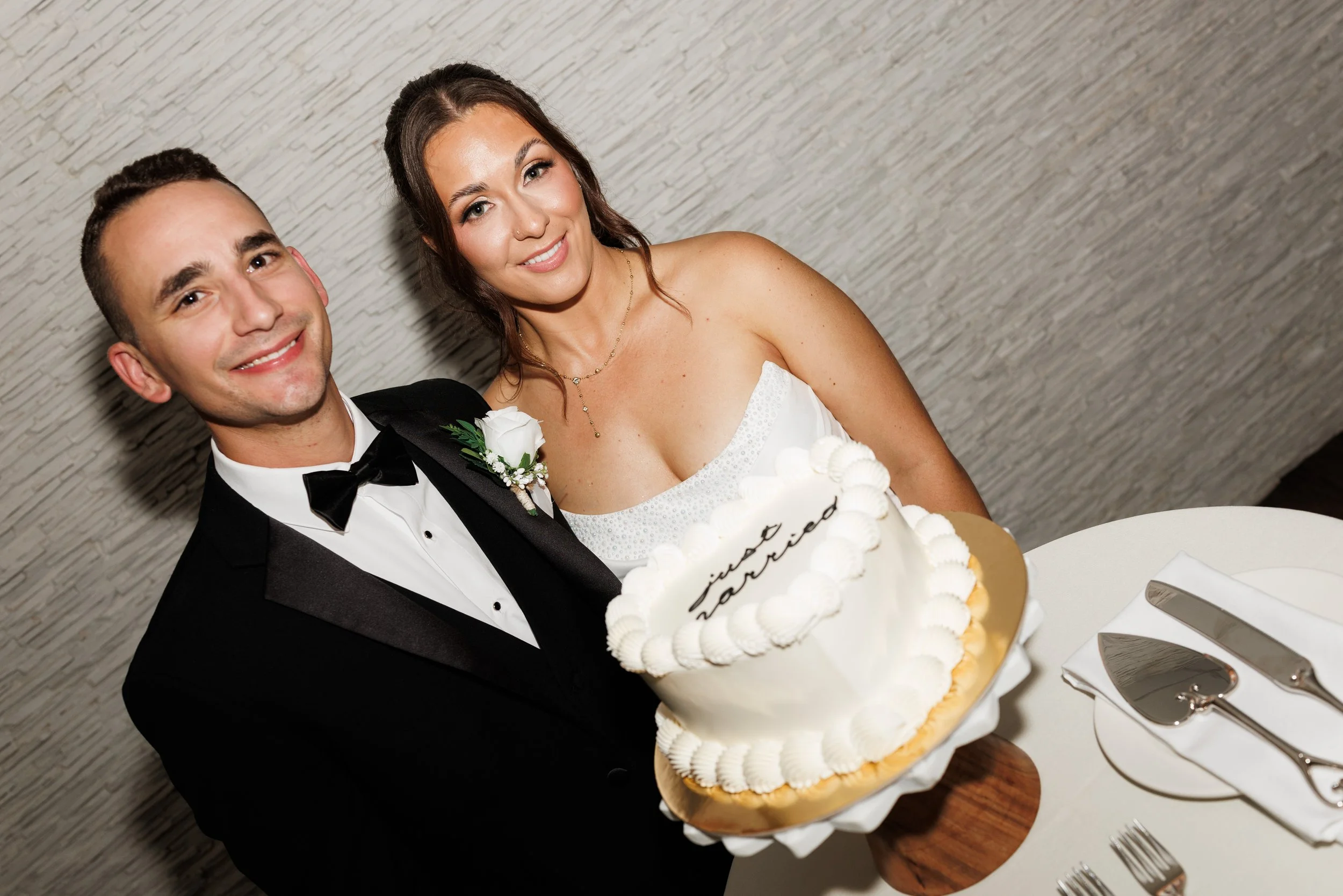 A groom and bride smiling at a wedding reception, standing behind a white, decorated wedding cake on a table, with utensils nearby.