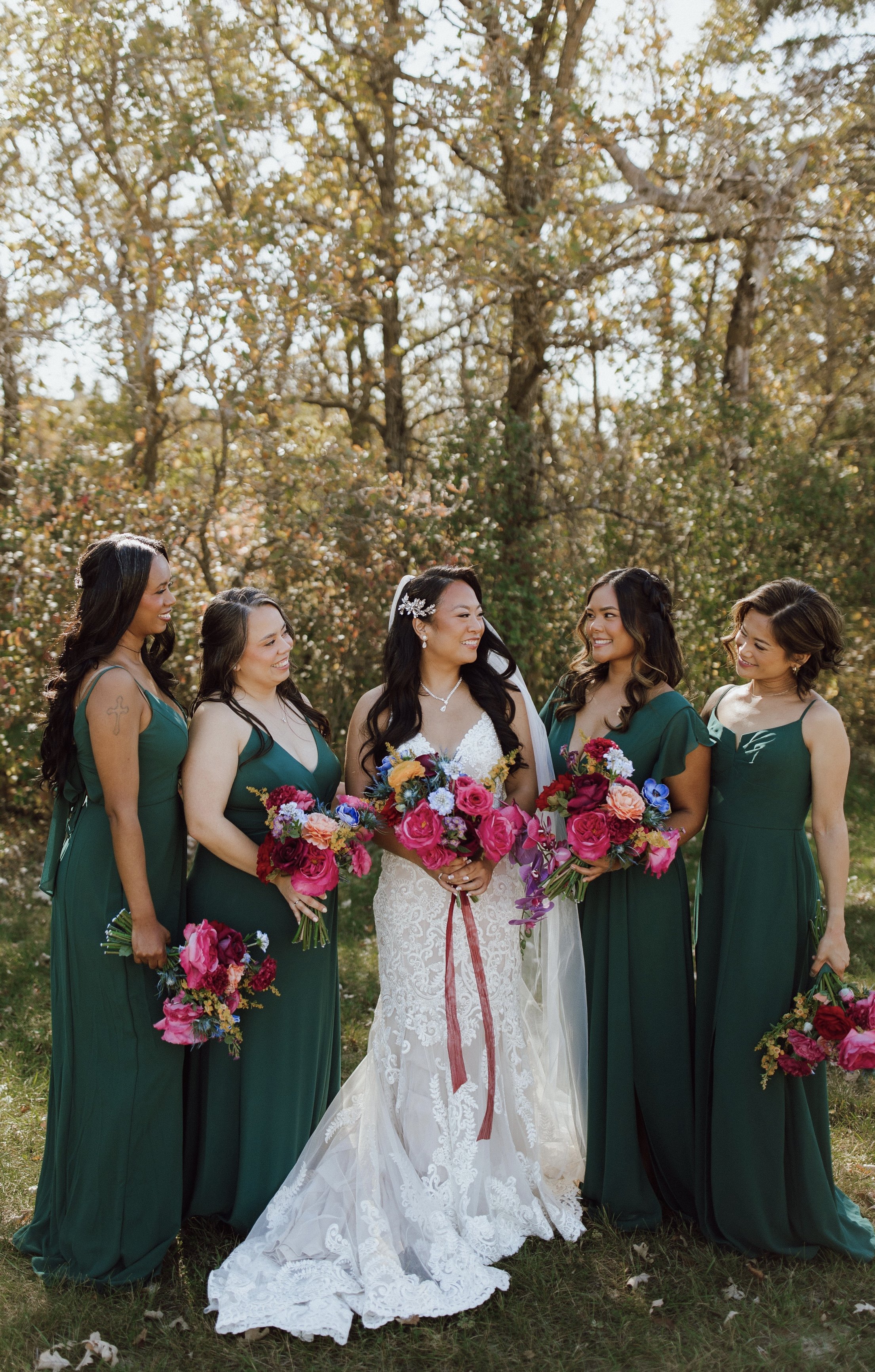A bride dressed in a white lace wedding gown holds a bouquet of colorful flowers, standing with five bridesmaids in matching green dresses outdoors under trees with autumn foliage, smiling and interacting.