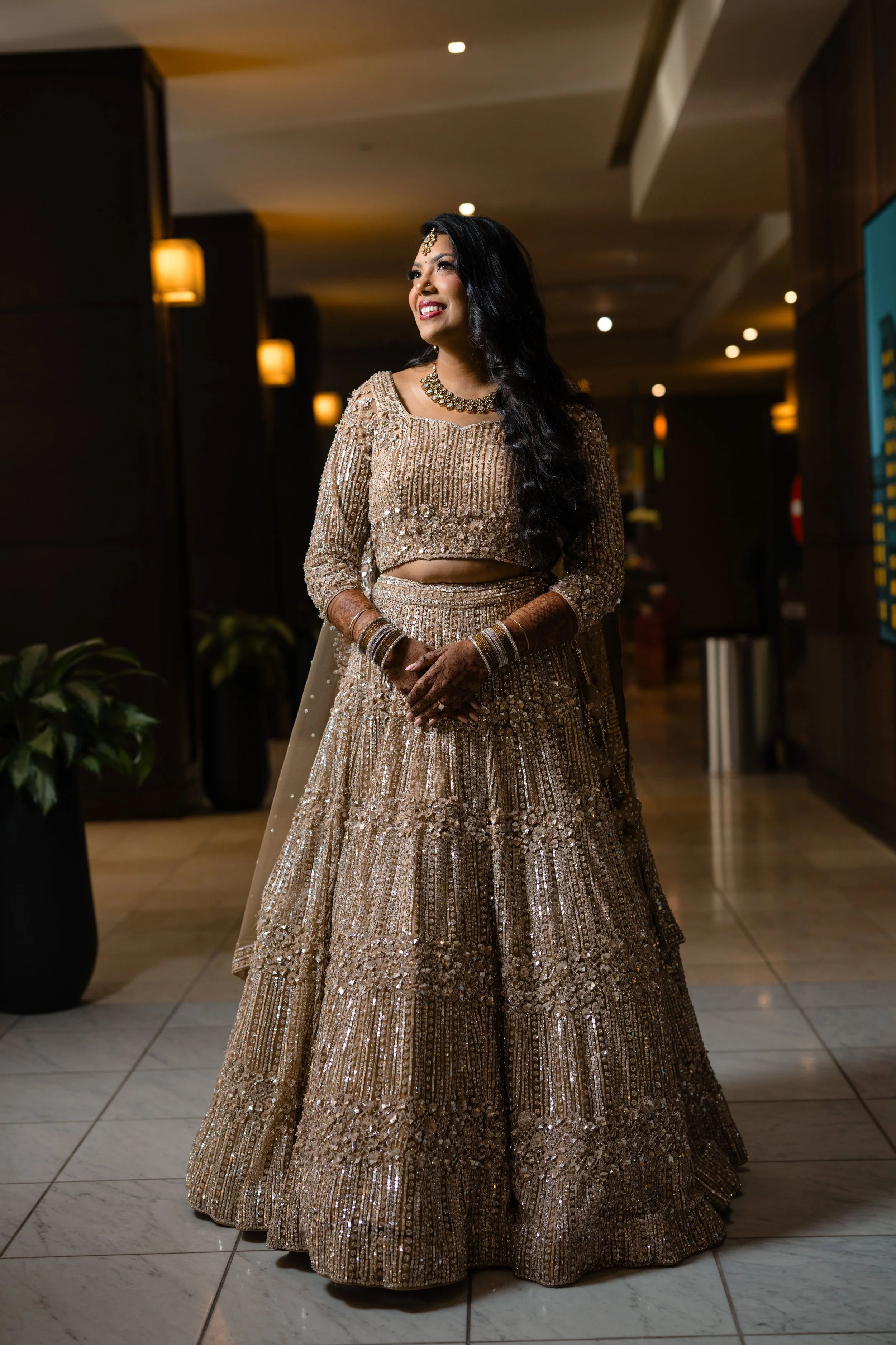 A woman wearing a traditional Indian bridal outfit, including a heavily embroidered gold and beige lehenga, matching jewelry and mehndi on her hands, standing indoors in a well-lit area with dark walls and wall-mounted lights.