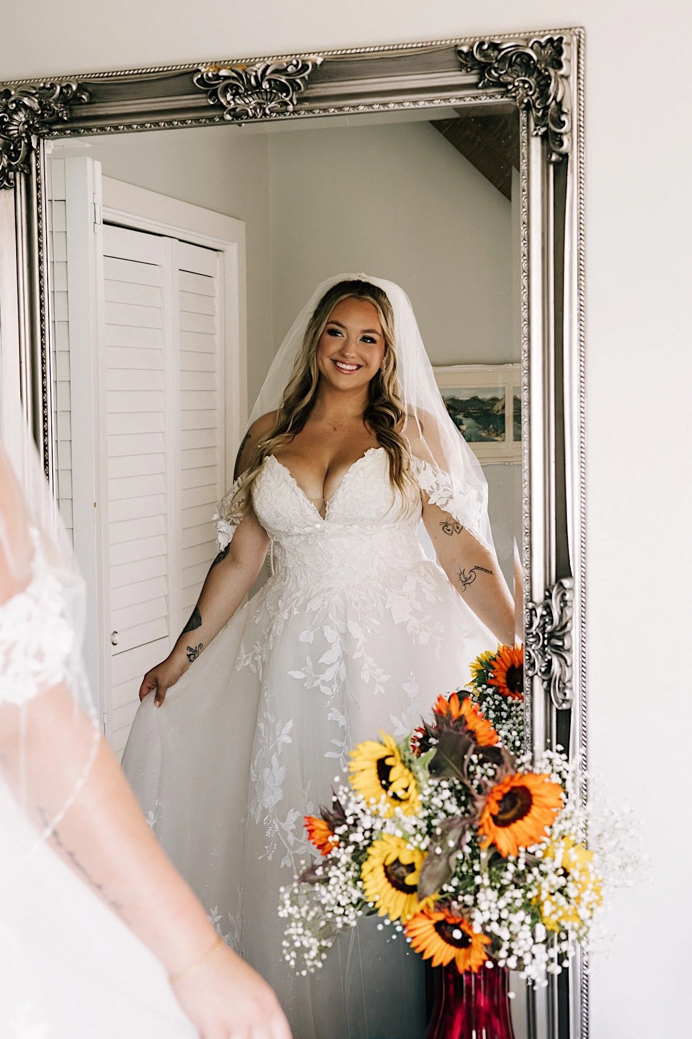 A happy bride in a white wedding dress looking at herself in a large ornate mirror, with a bouquet of sunflowers and other flowers in front of the mirror.