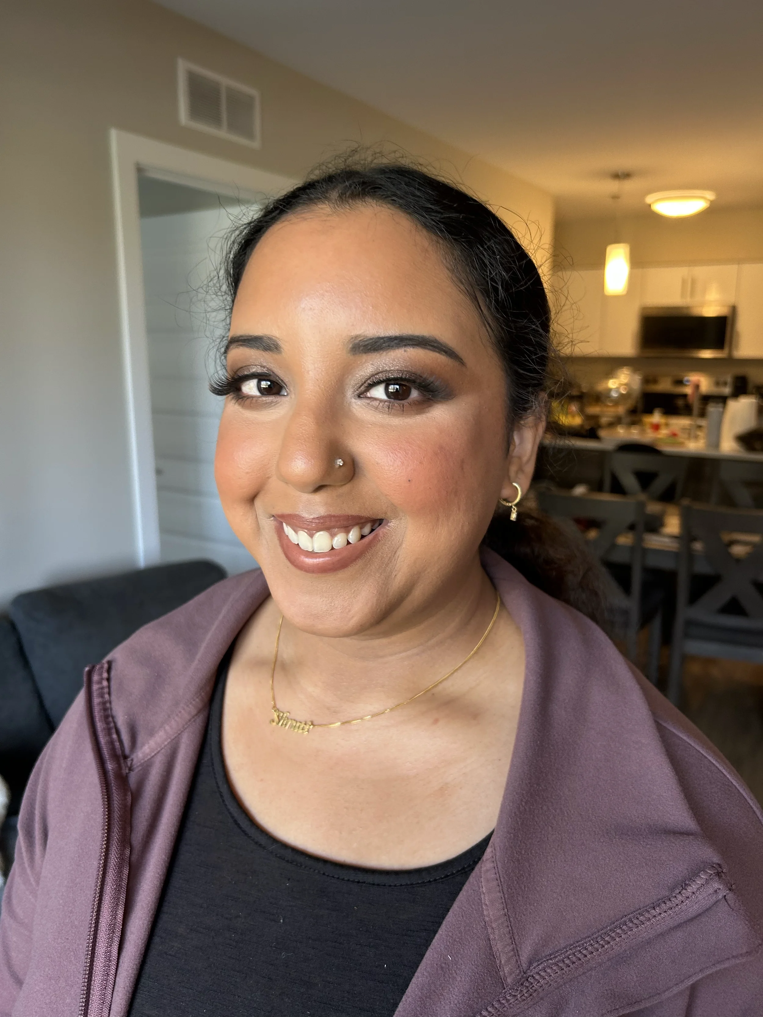 Close-up of a woman smiling in a cozy indoor setting, wearing a dark top, purple hoodie, gold necklace, and earrings, with a blurred background of a kitchen and dining area.