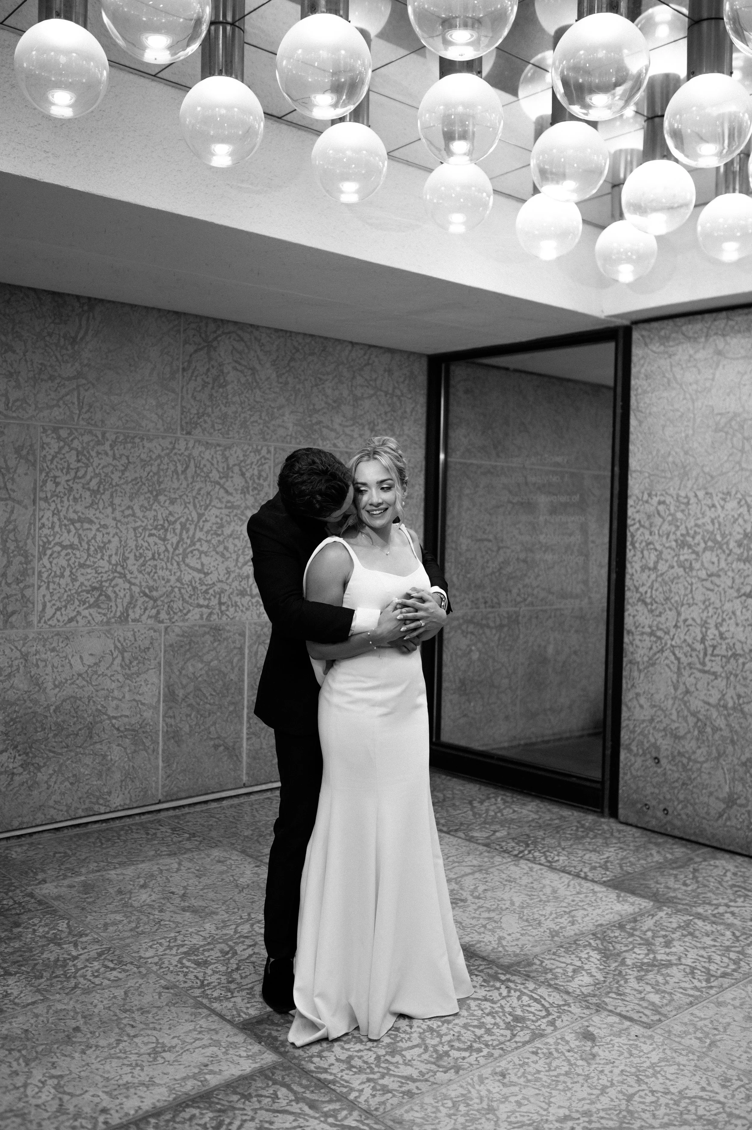 A black-and-white photo of a man and woman dancing closely at a formal event. The woman is wearing a white gown and the man is in a black tuxedo. They are smiling and holding each other, with the woman's hands clasped together in front of her, in a room with patterned carpet, textured walls, and overhead spherical light fixtures.