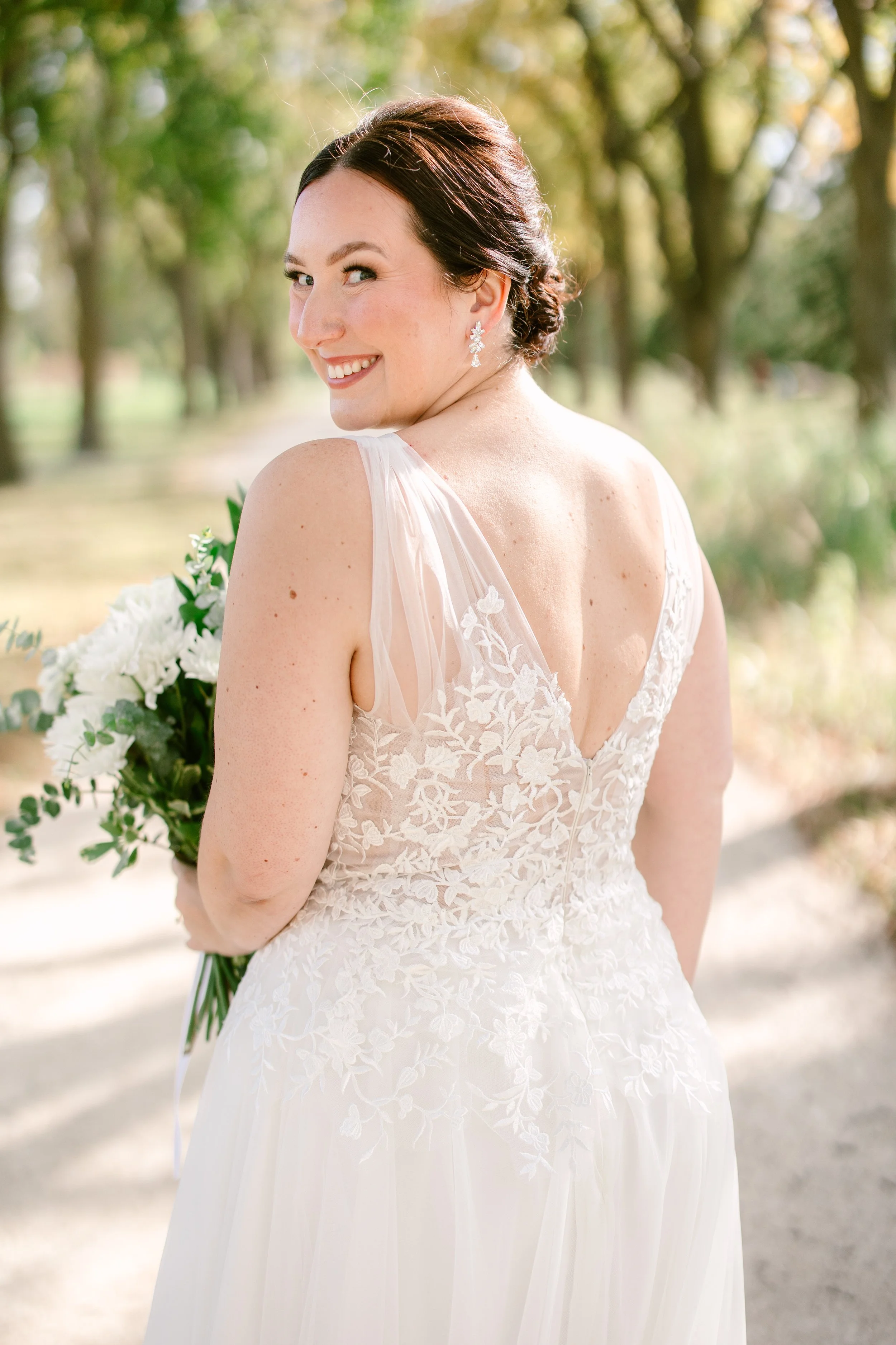 A bride outdoors with trees in the background, wearing a lace wedding dress and floral earrings, holding a bouquet of white flowers, smiling and looking over her shoulder.