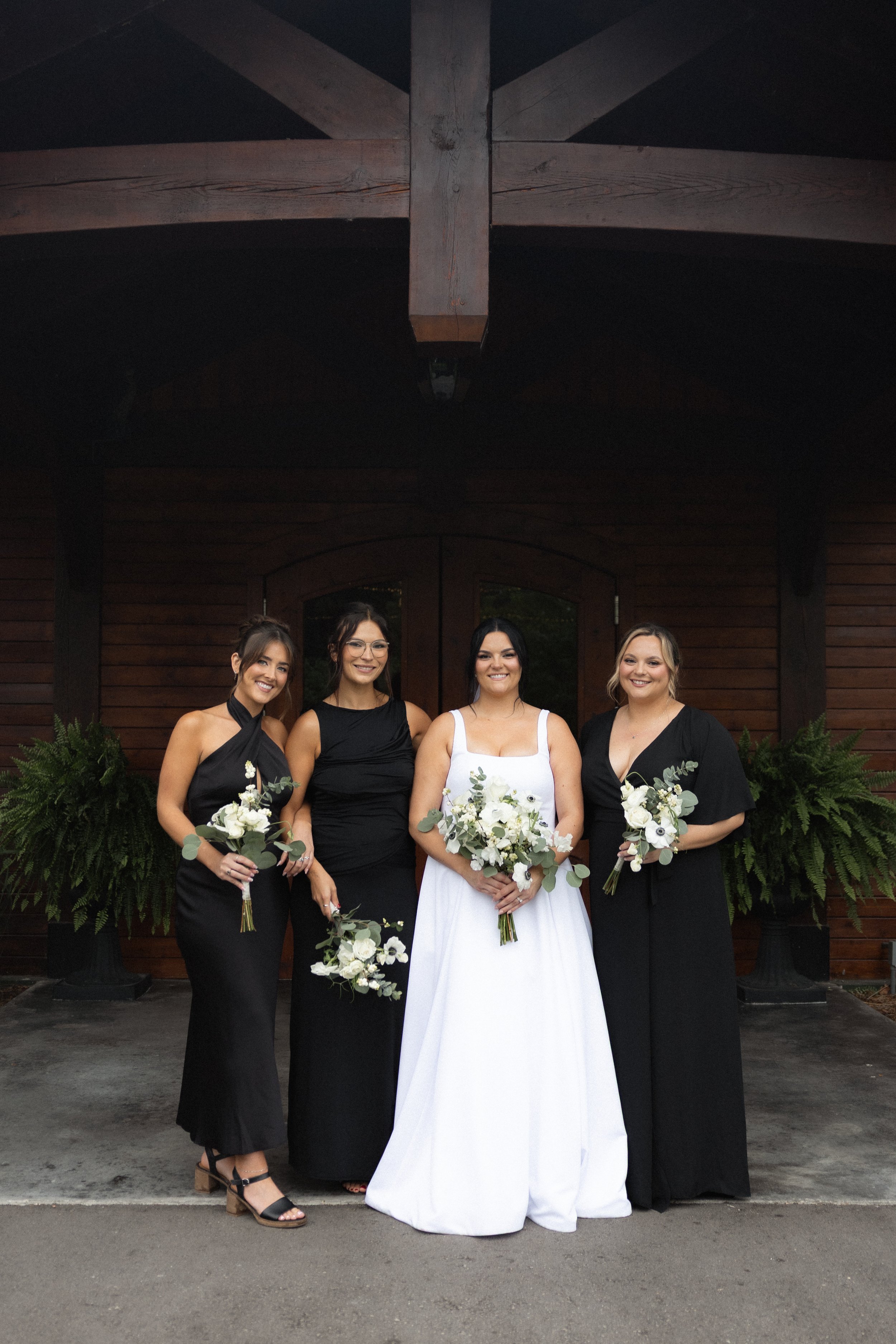 Four women in formal attire standing outdoors in front of a wooden building with large doors, holding white floral bouquets, smiling at the camera. The woman in the middle is wearing a white dress, and the other three wear black dresses.