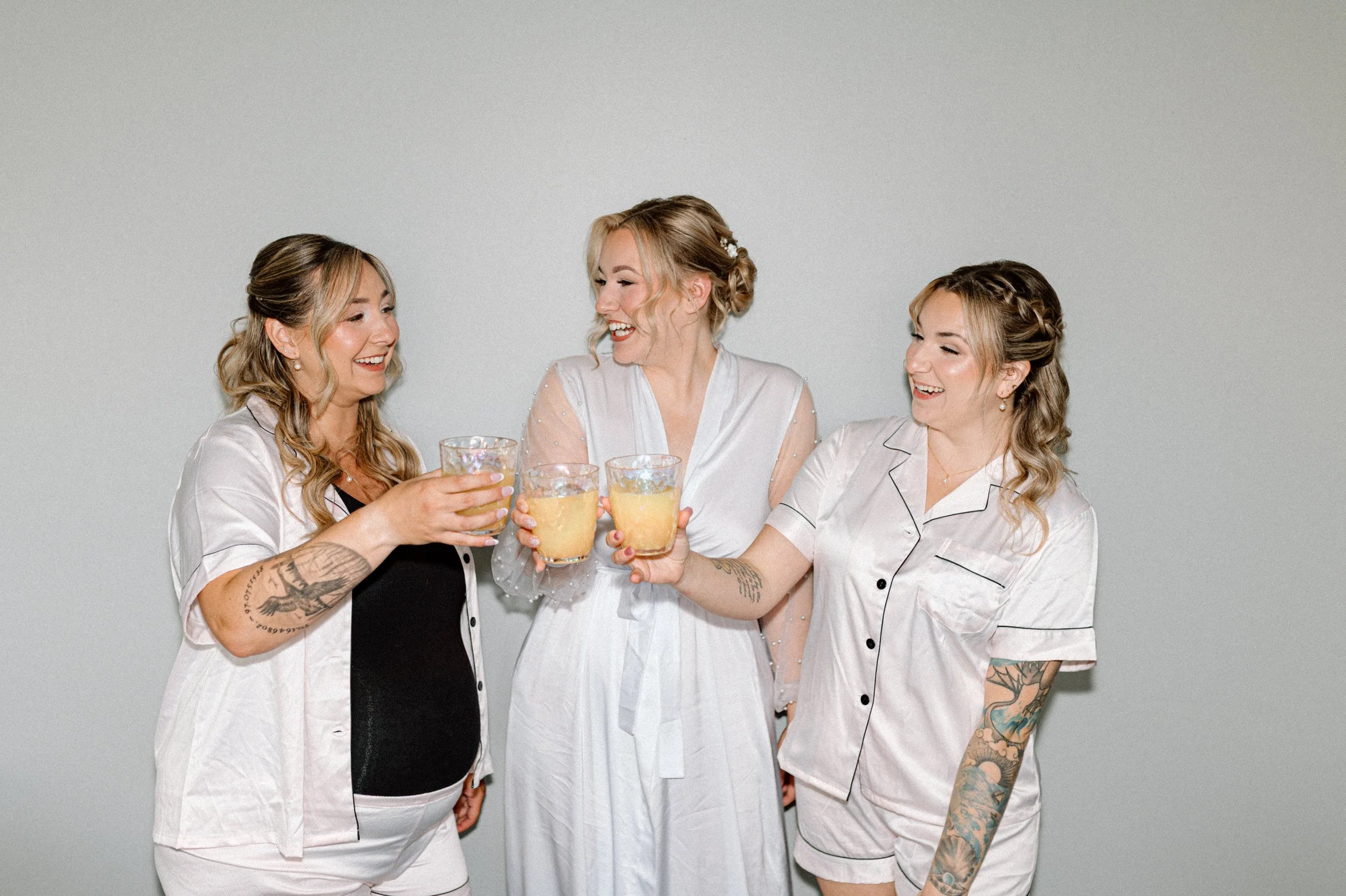 Three women in pajamas smiling and raising glasses for a toast in front of a plain light gray background.