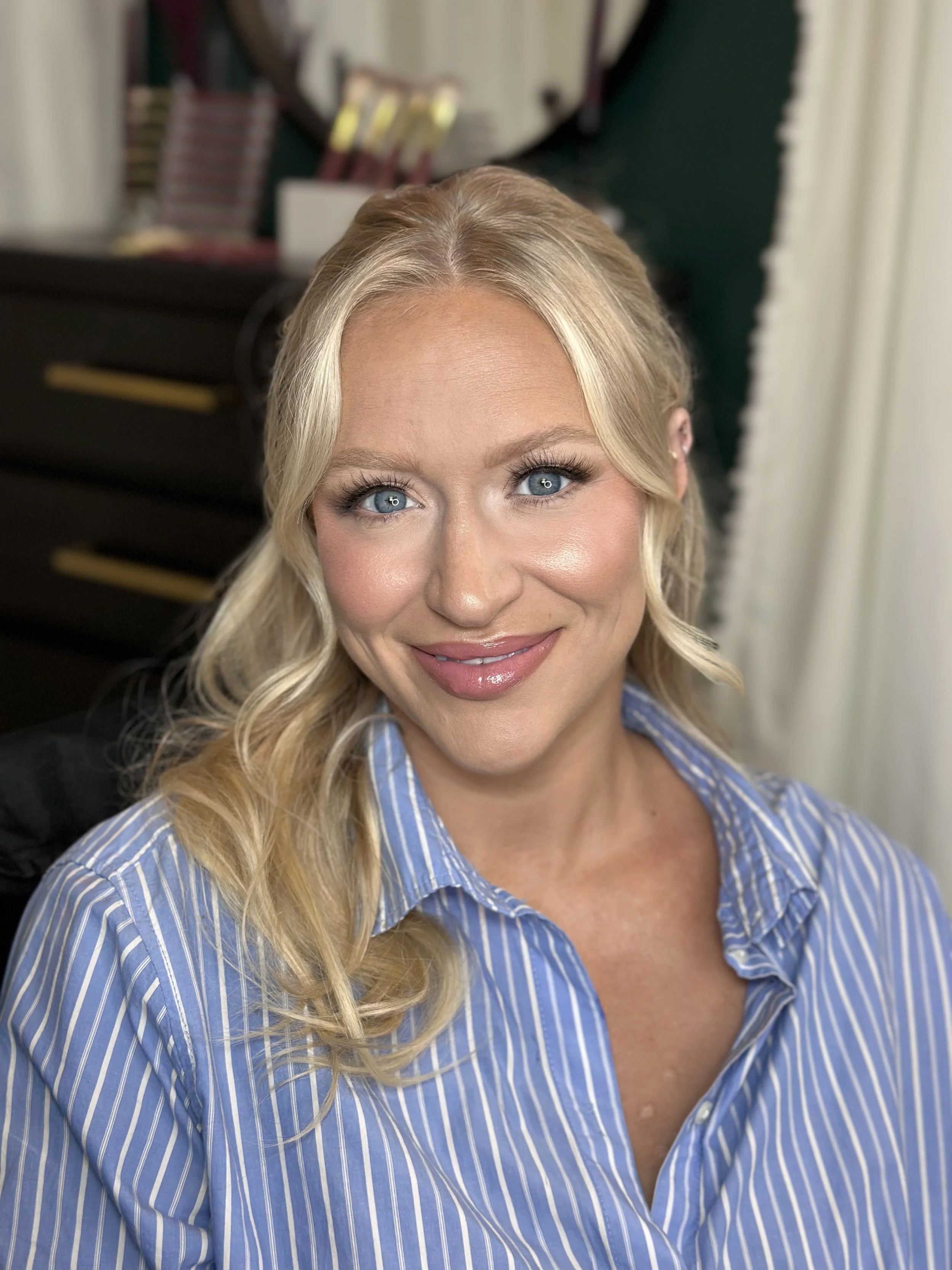 A woman with long, wavy blonde hair, blue eyes, and light makeup wearing a blue and white striped shirt, smiling at the camera.