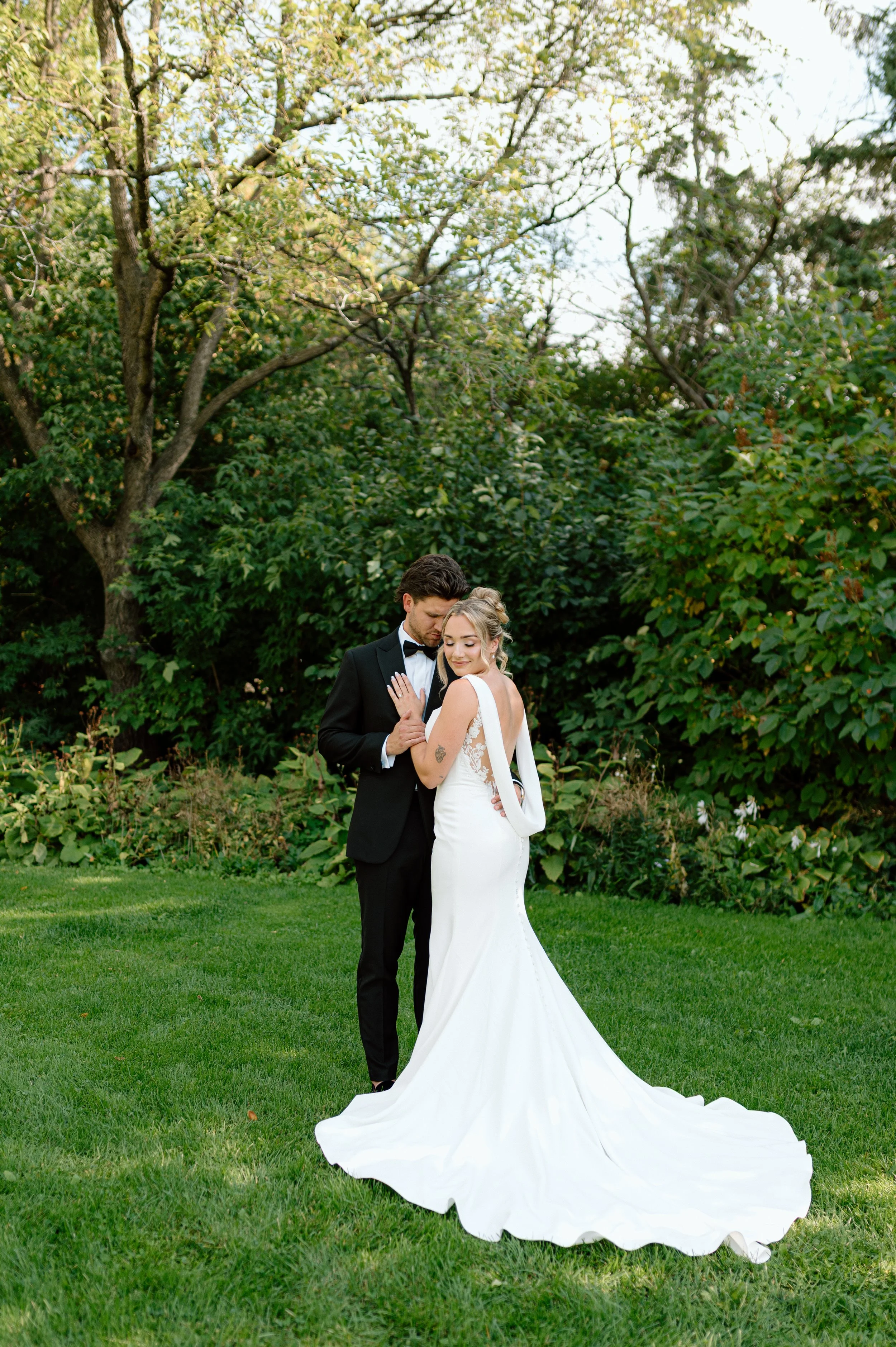 A bride and groom standing closely in a garden, with the groom in a tuxedo and the bride in a white wedding dress, surrounded by greenery and trees.