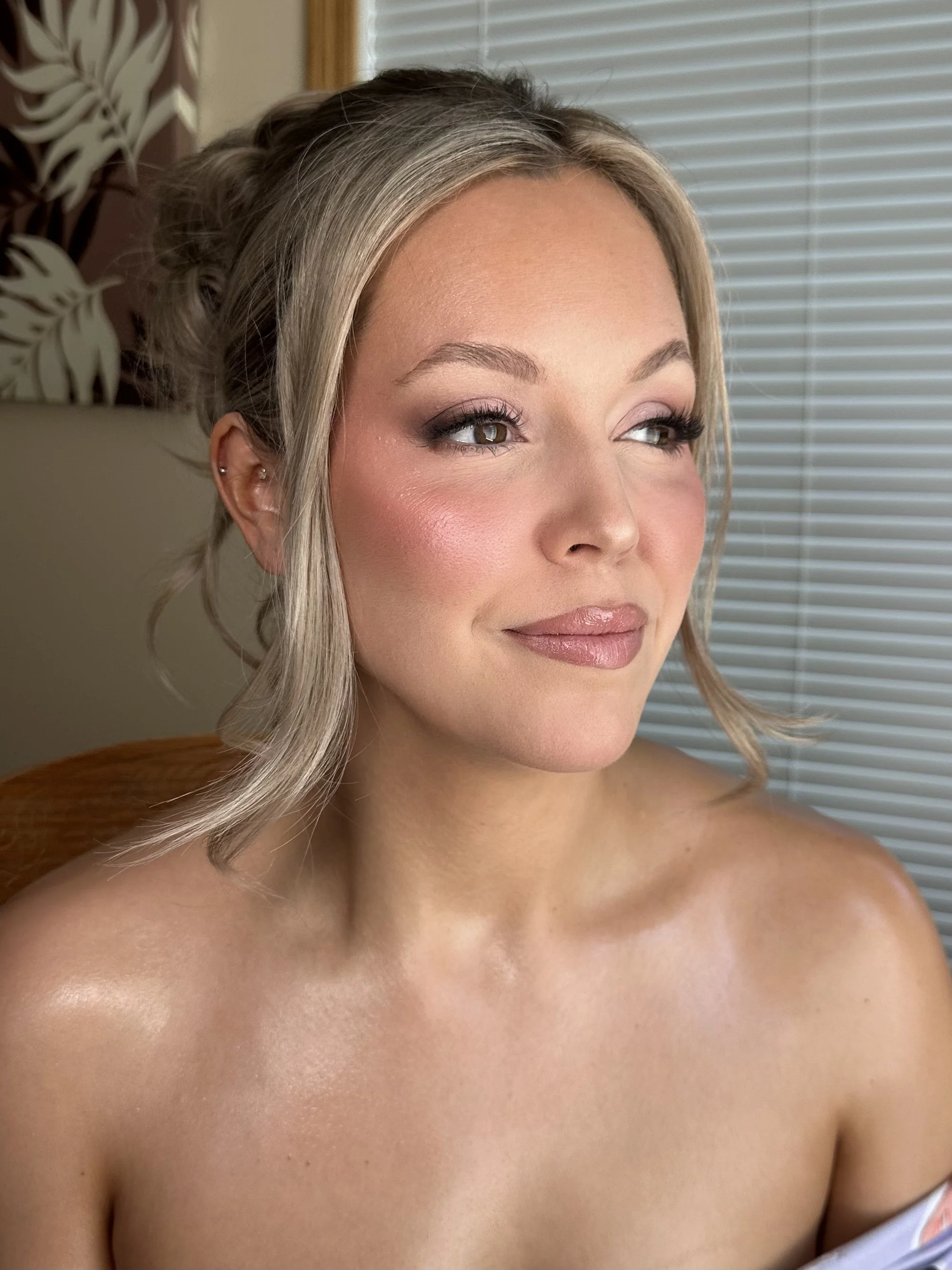Close-up of a woman with blonde hair styled in loose waves, wearing makeup, looking off to the side with a slight smile, in front of a window with blinds.