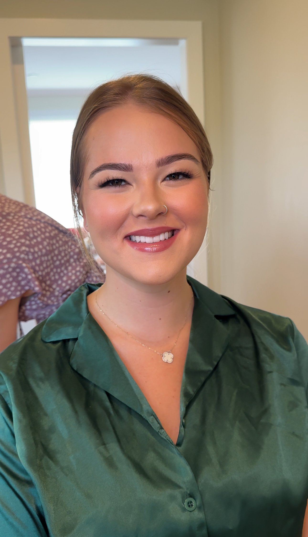 Close-up of a woman smiling with light makeup, wearing a green satin blouse, a delicate necklace, and a nose ring, in a well-lit indoor setting.