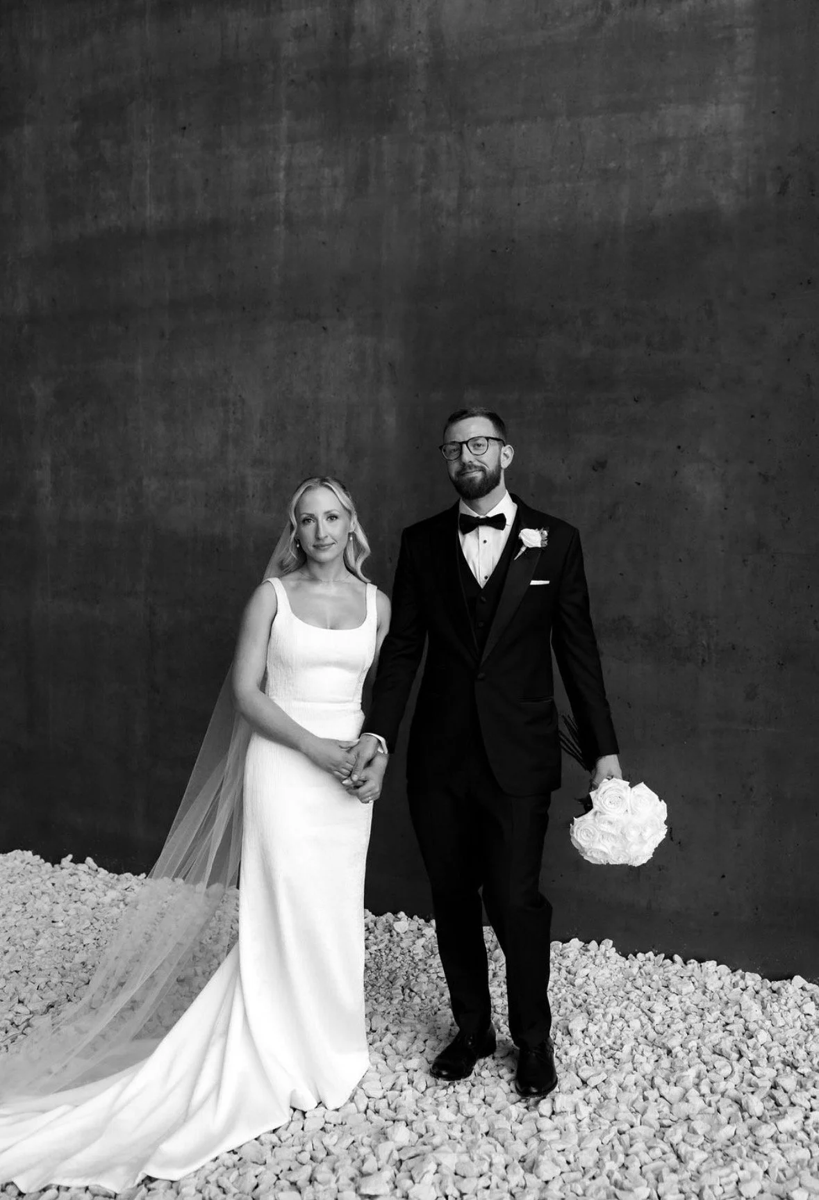 A bride and groom holding hands, standing on rocky ground against a plain wall, dressed in wedding attire, with the groom holding a bouquet of white roses.