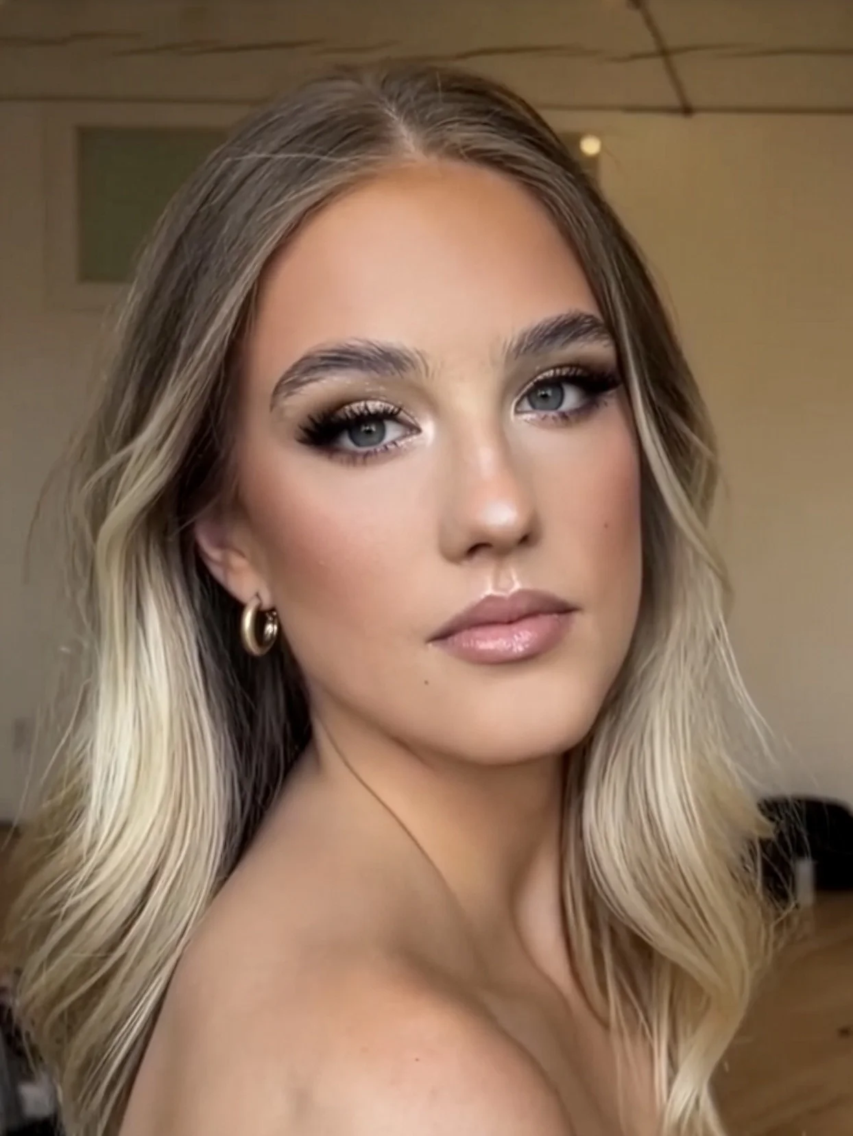 A woman with long blonde hair and blue eyes, wearing makeup with mascara, eyeliner, and nude lipstick, and accessorized with gold hoop earrings, looking slightly to the side against an indoor background.