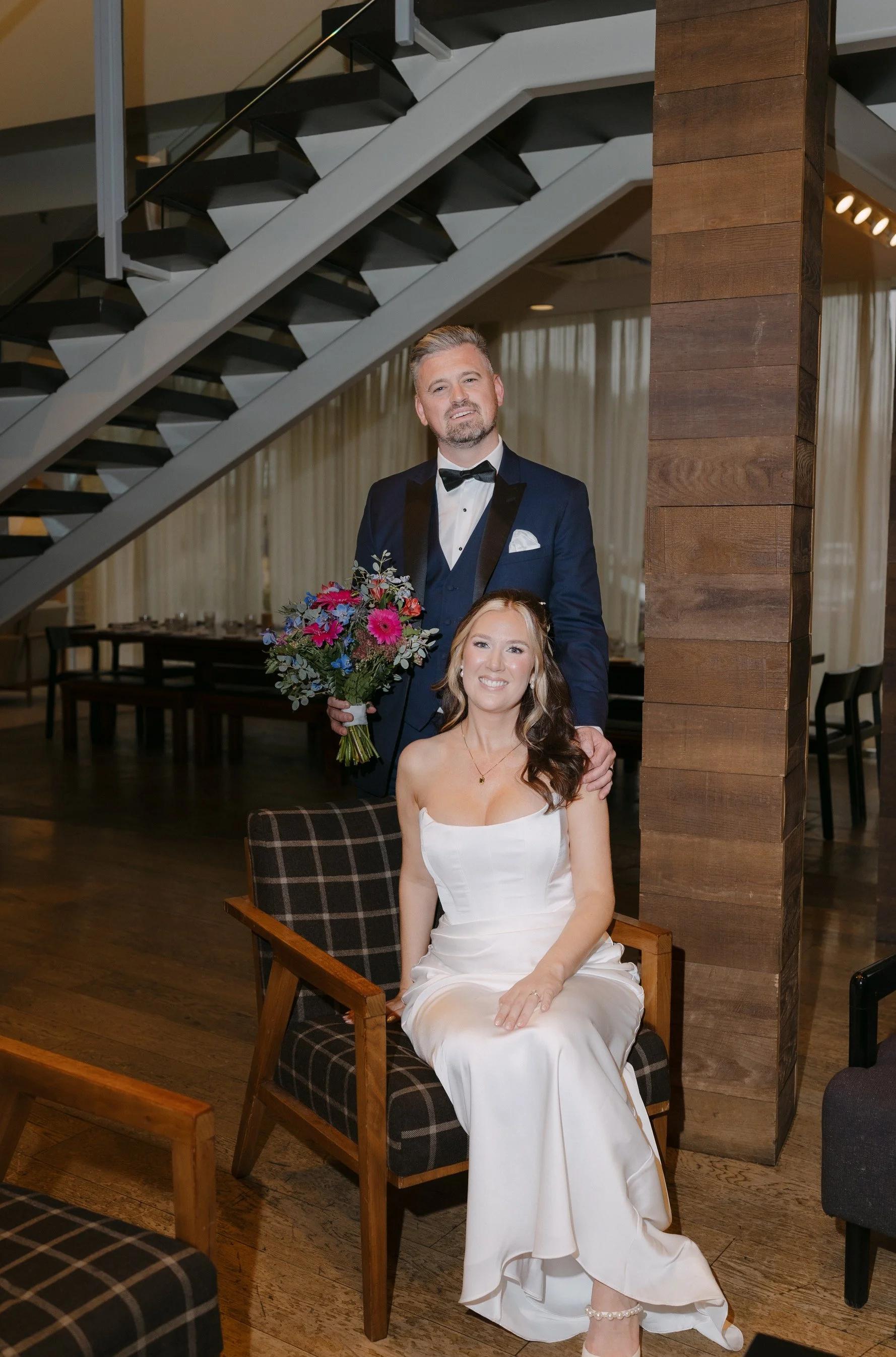 A man in a tuxedo holding a bouquet of flowers standing behind a seated woman in a white dress in a warmly lit room with wood accents and a staircase.
