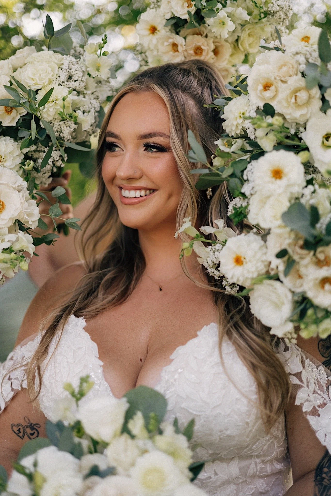 A woman in a white lace wedding dress surrounded by a large arrangement of white flowers and greenery, smiling and looking to the side.