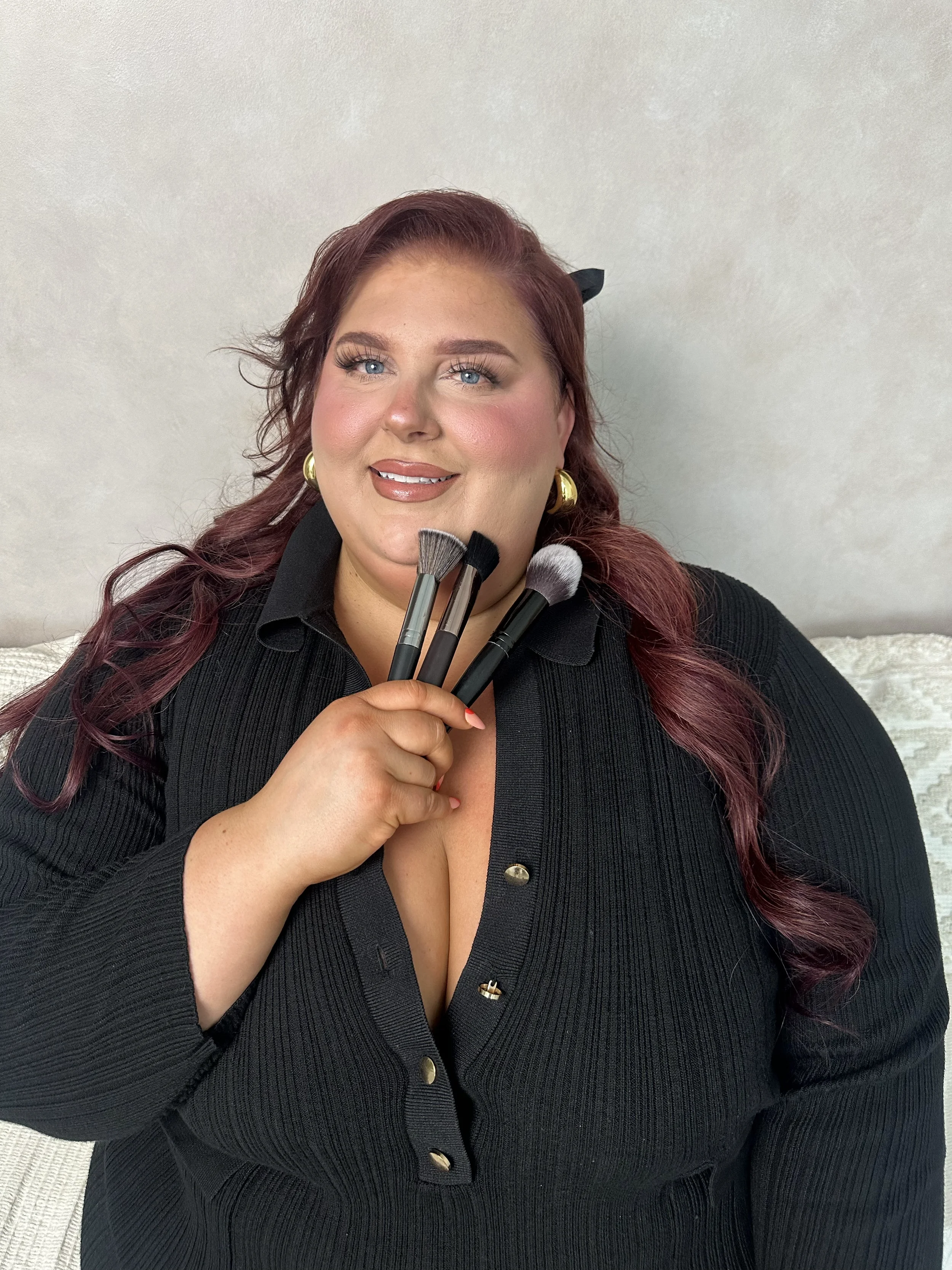A woman with red hair and blue eyes holding makeup brushes, wearing a black top with buttons, gold earrings, and smiling, against a plain light-colored wall.