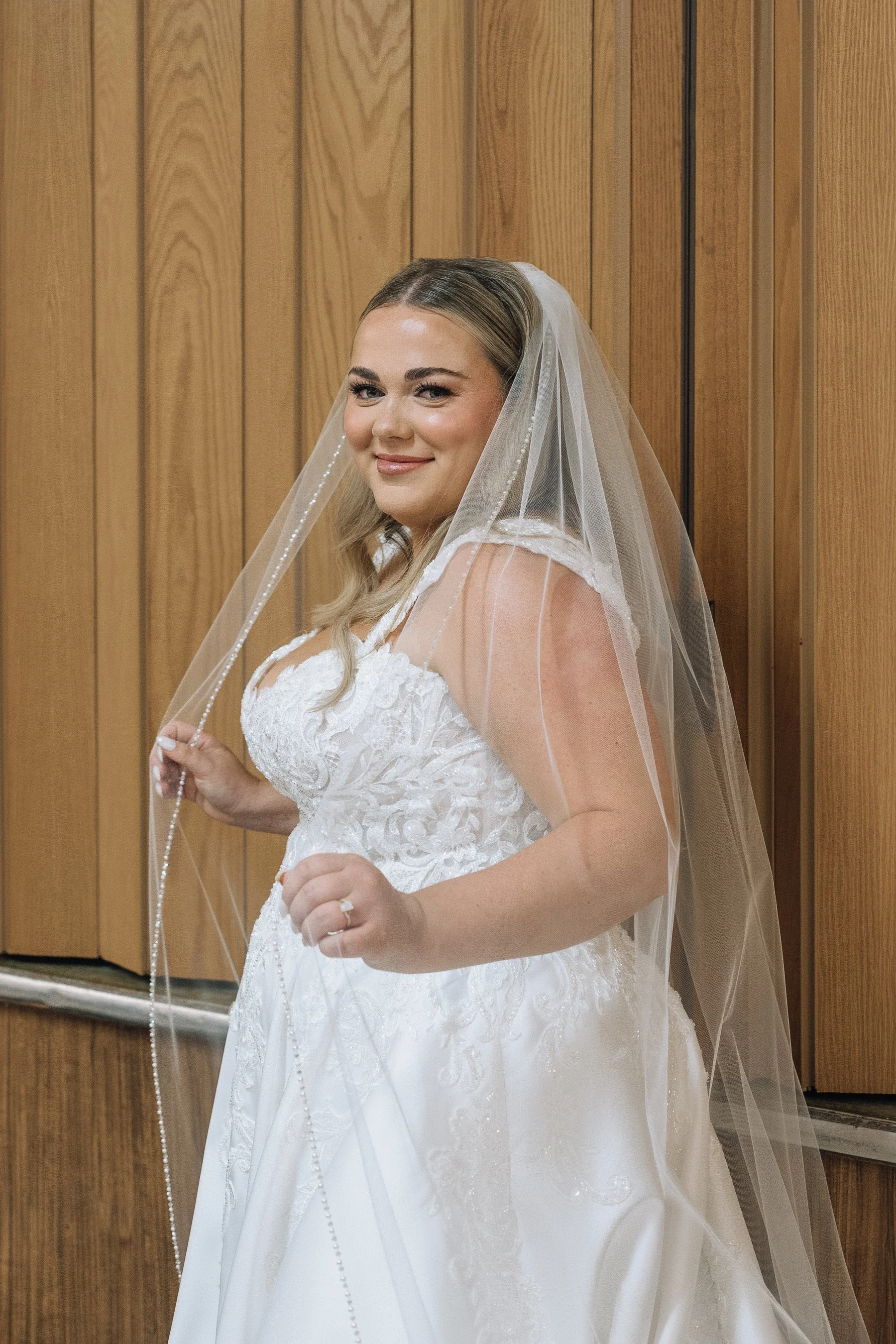 A smiling bride in a white wedding dress with lace details, holding a pearl necklace, standing in front of a wooden panel wall, wearing a veil.