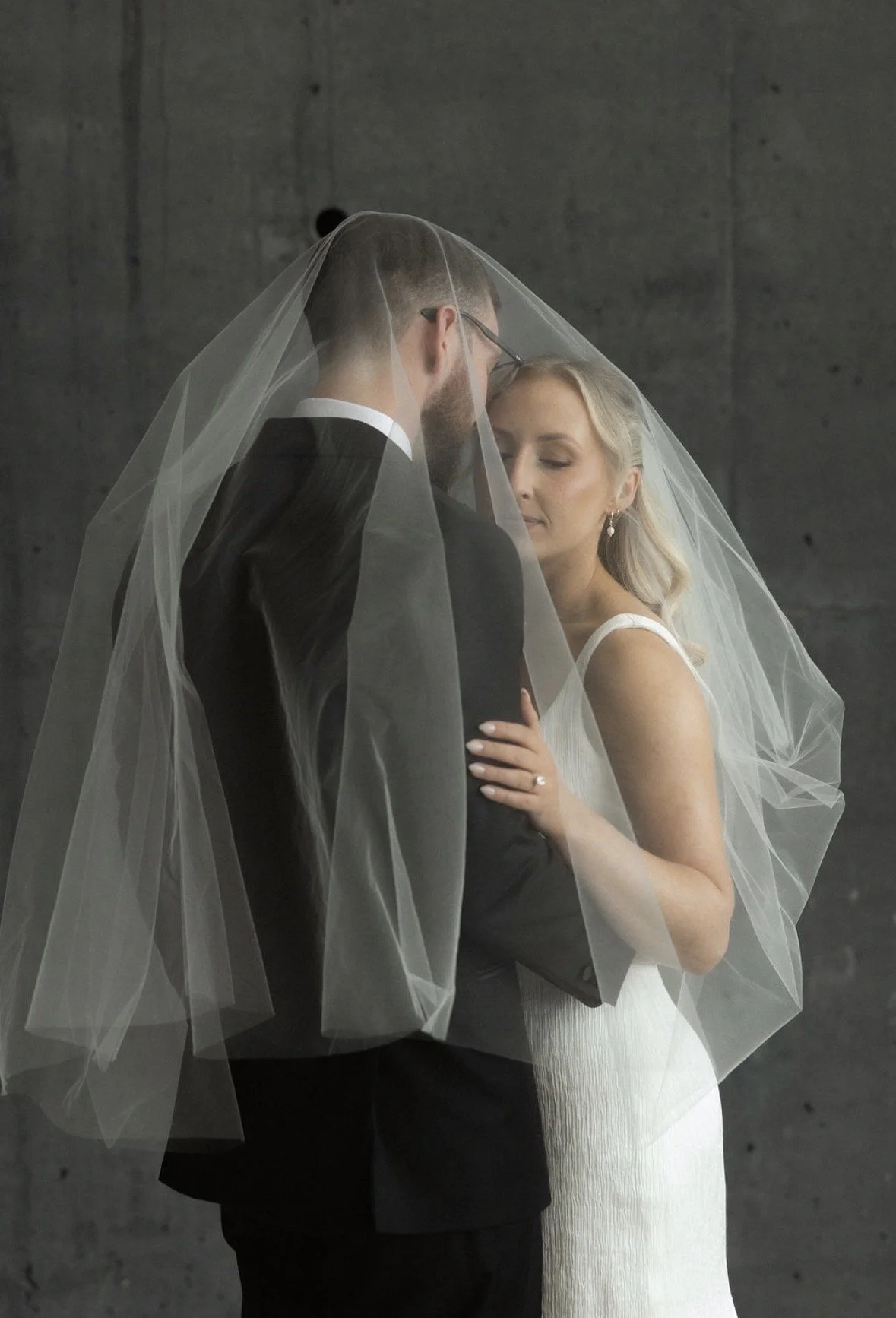 A bride and groom standing close together, with the bride gently touching the groom's chest. The groom is wearing a black tuxedo with a white shirt, and the bride is in a white dress with a veil covering both of their heads. They are facing each othe