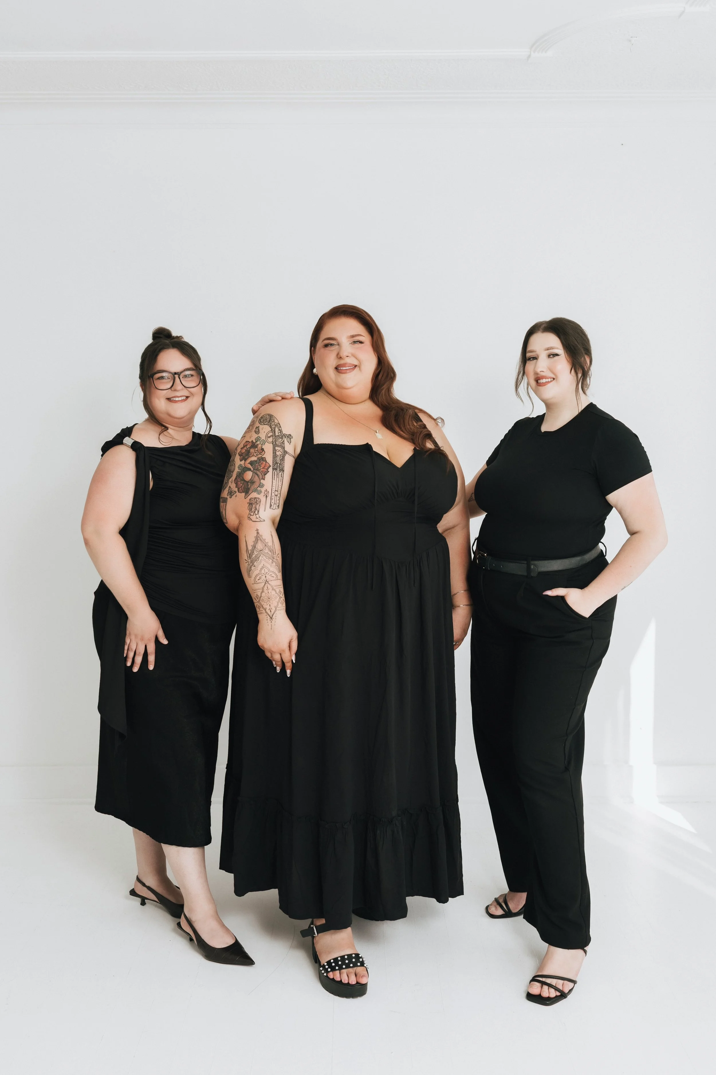 Three women standing together in a white room, all dressed in black outfits, smiling at the camera.