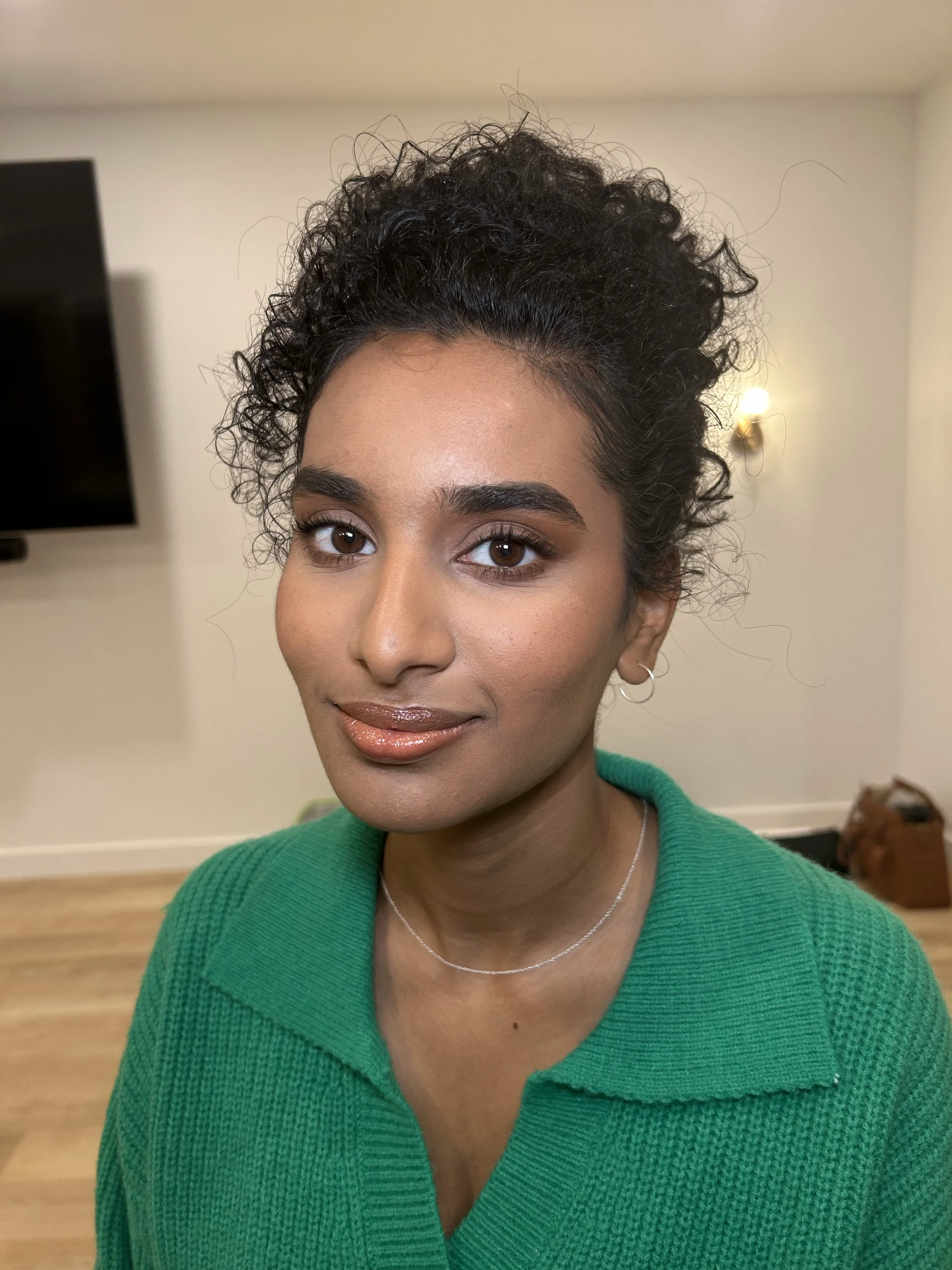 Close-up of a woman with curly black hair, wearing a green sweater, silver earrings, and a silver necklace, standing indoors.