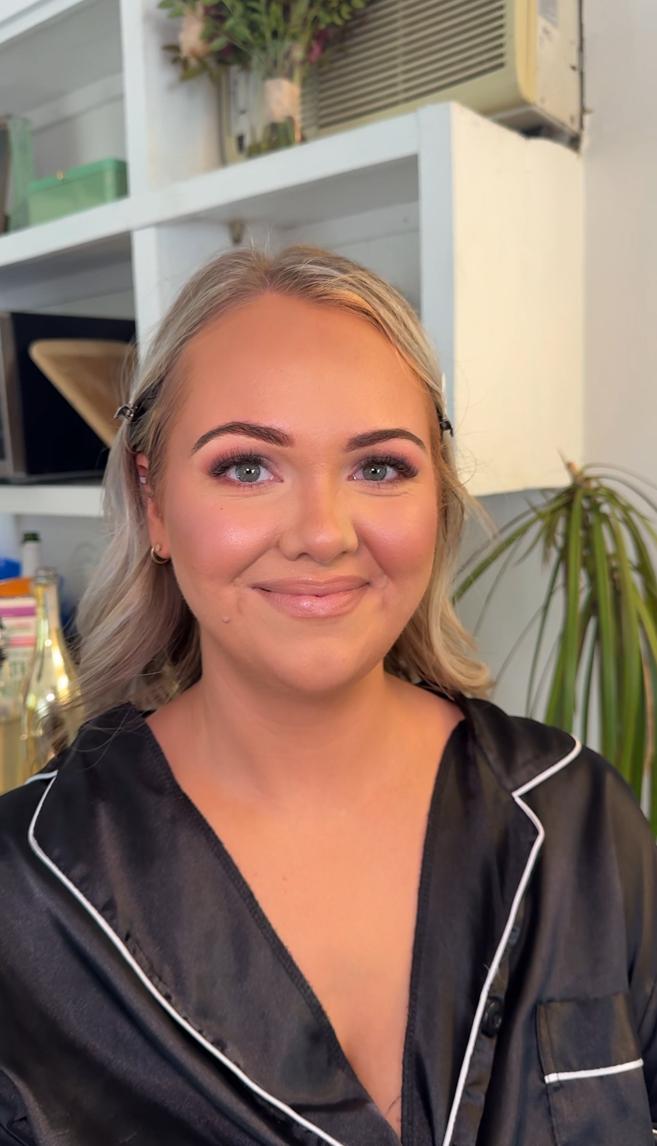 A woman with blonde hair and makeup, wearing a black satin outfit with white piping, smiles at the camera inside a room with shelves, plants, and an air conditioning unit in the background.